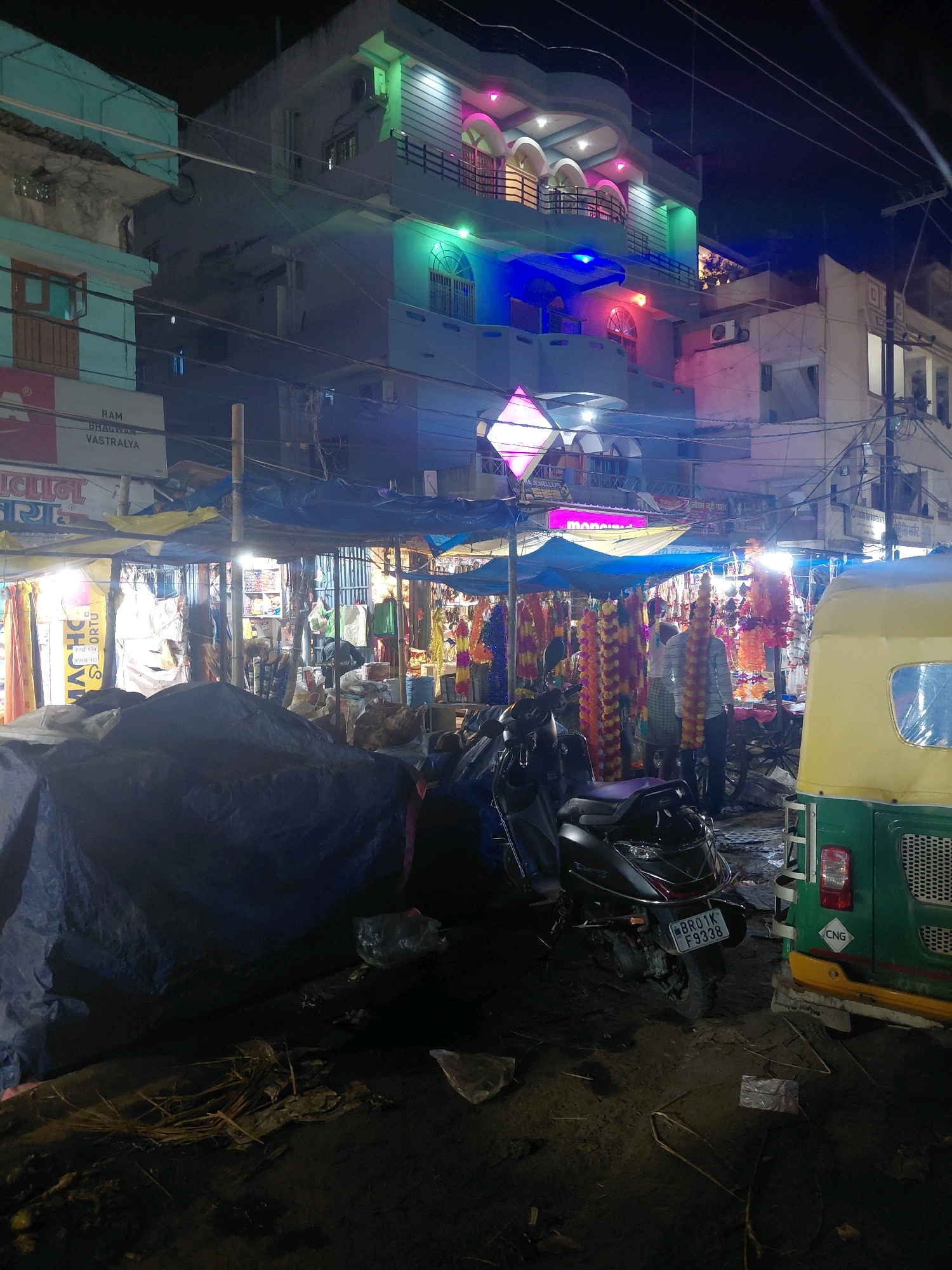 An outdoor market scene at night in Patna, India, is depicted, with a focus on a street vendor's stall. The stall is covered by blue and yellow tarps, with a variety of goods, including colorful garlands of flowers, displayed. Two men are visible within the market area; one is standing and wearing a striped shirt and checkered lungi, with garlands draped around his neck. Another man, wearing a blue checkered shirt and lungi, is partially obscured.
To the right of the stall, a green and yellow auto-rickshaw is parked. In the foreground, several scooters are parked, one of which has a license plate that reads "BR01K F9338". The ground appears to be dirt and is littered with debris, including plastic bags and dry vegetation. In the background, a multi-story building is visible, illuminated with colorful lights, including pink, green, and blue. Several signs are also present, one reading "RAM BHAGWAN VASTRALYA". The overall impression is a bustling, albeit somewhat cluttered, night market.
