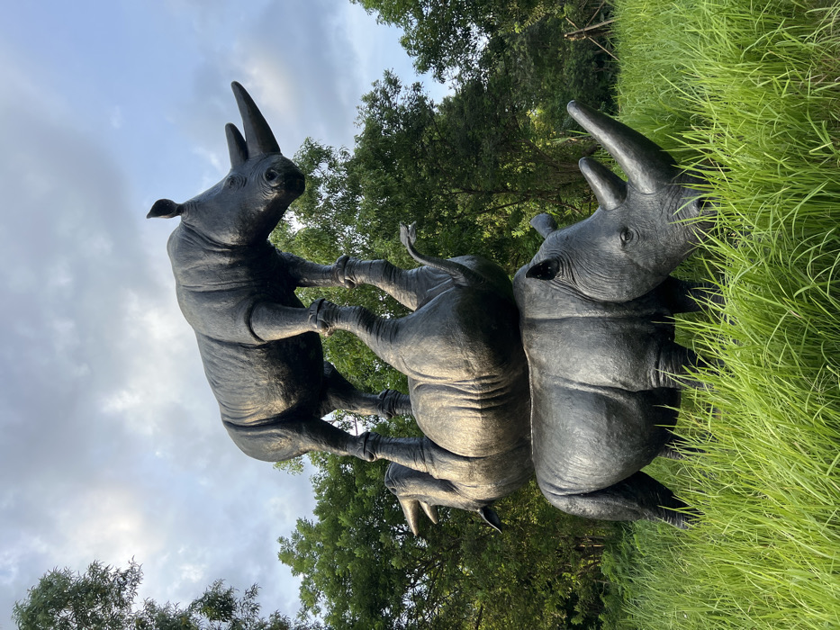A large-scale public art installation is depicted outdoors in Pecatu, Indonesia. The central subject is a stack of three dark, metallic rhinoceros sculptures. The lowest sculpture stands upright on all four legs, partially obscured by tall, vibrant green grass, with its head angled downwards as if grazing. Resting on the back of this bottom rhinoceros is a second sculpture, positioned upside down and balanced on its back, with its four legs extending upwards. The third and uppermost rhinoceros sculpture stands upright on all four legs atop the inverted middle rhino, with its head also slightly lowered. The background consists of dense green foliage from numerous trees, extending to a partly cloudy sky. No human figures are present. The lighting is soft, indicative of either early morning or late afternoon.