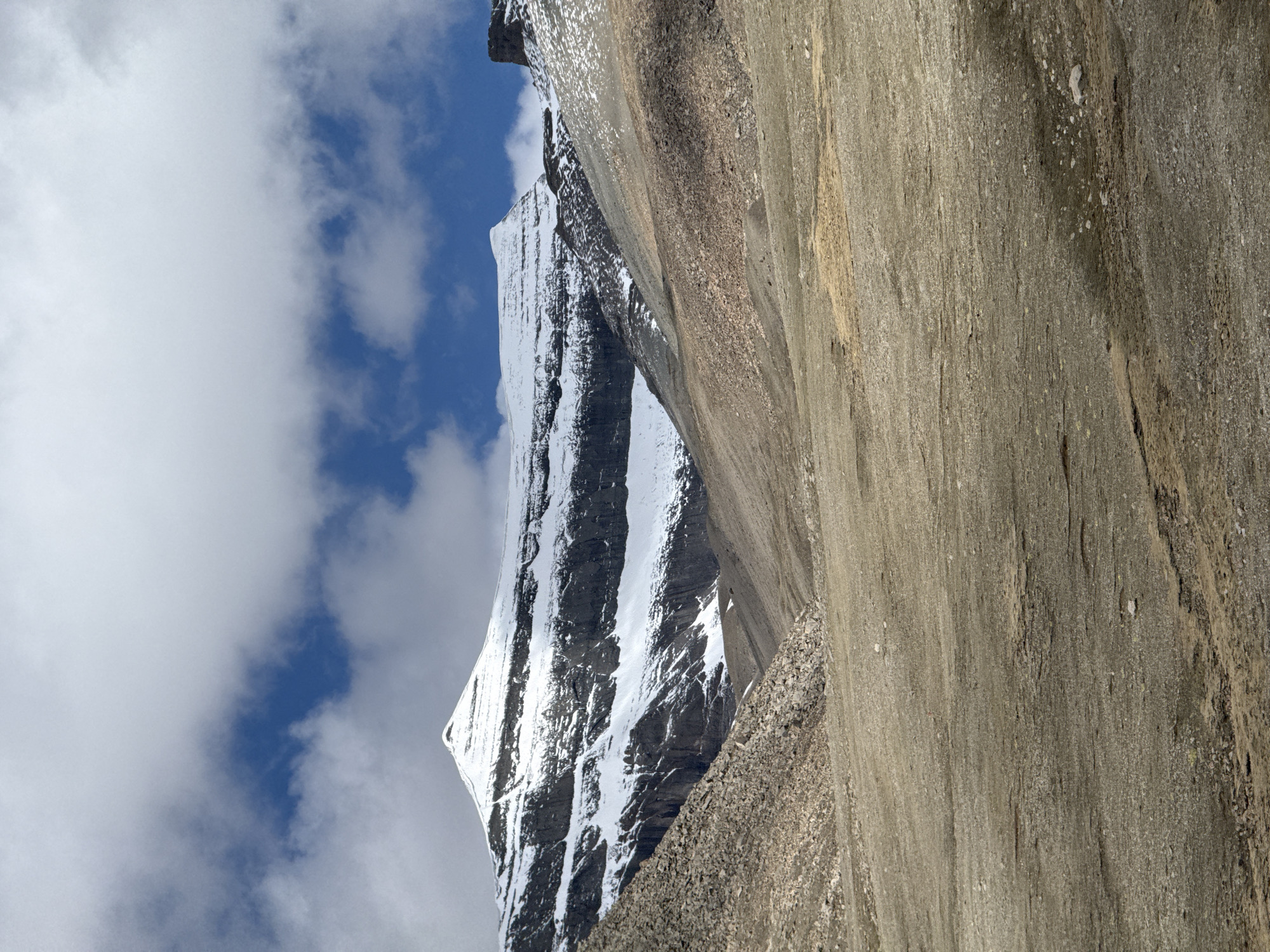 The image depicts a dramatic, high-altitude mountain landscape under a partially cloudy blue sky. A towering, snow-capped mountain range dominates the left side of the frame, its jagged peaks and steep slopes covered in brilliant white snow that contrasts with dark, rugged rock faces. Vertical streaks and patches of snow and ice are visible on the mountain's sides, reflecting bright sunlight. The top of the main snow-capped peak is sharply defined against the sky.
In the foreground, a massive, textured rock face or cliff occupies the right side of the image. This rock surface displays varying shades of grey and earthy brown, with prominent vertical striations and a granular, rough texture. Small, scattered white and light-colored pebbles and debris are visible on this rock surface and at its base.
The sky overhead features patches of deep blue peeking through soft, white, and light-grey cumulus clouds. The strong, clear illumination indicates daytime, likely mid-morning or afternoon. There are no visible people, animals, man-made objects, or text. The scene conveys a sense of vastness, grandeur, and remote natural beauty in an alpine environment.