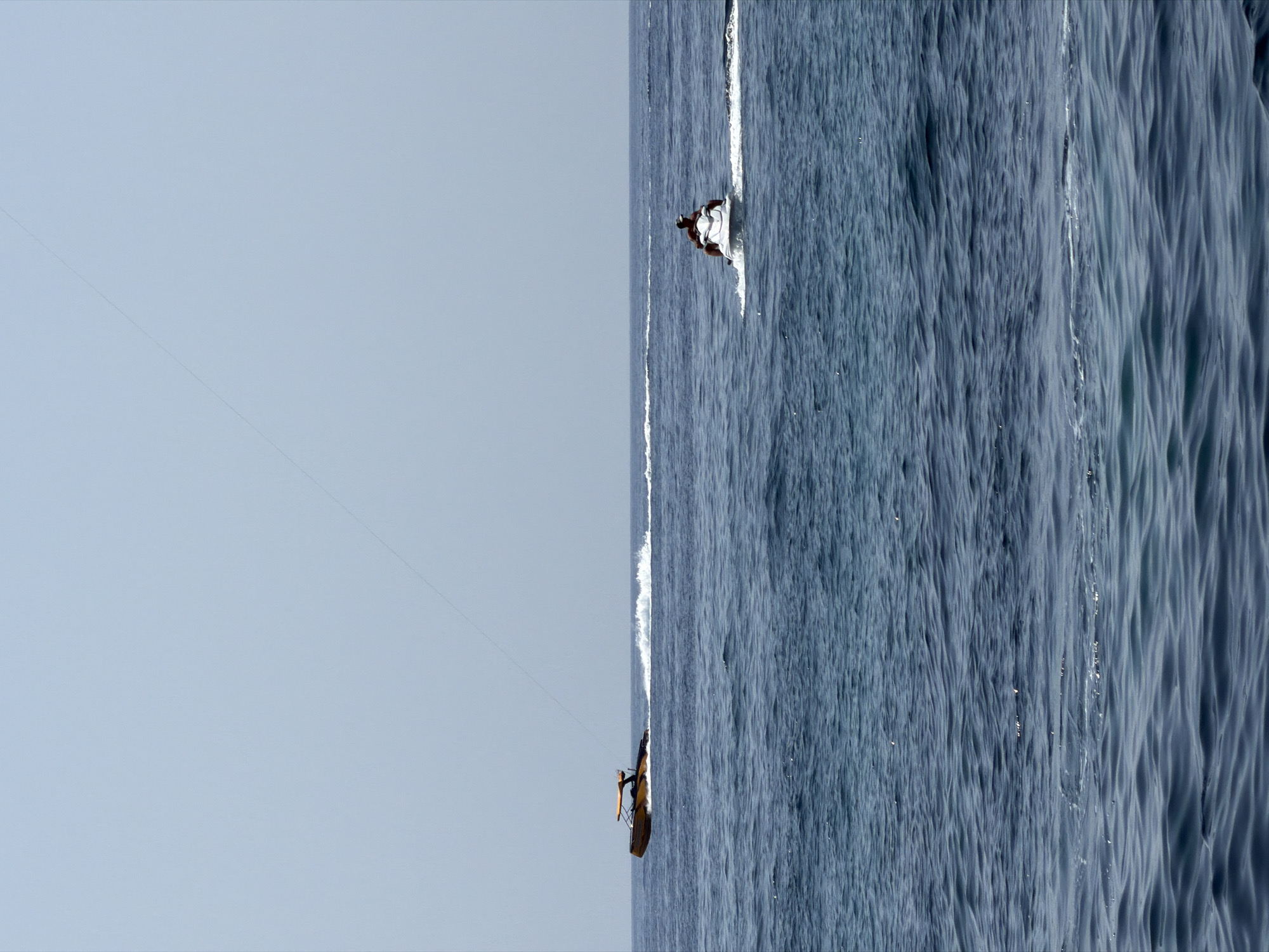 Two vessels are navigating the Adriatic Sea off the coast of Budva, Montenegro. In the mid-left, a small, orange and black motorboat is cutting through the water, leaving a white wake. A tow rope, likely for water sports, extends from the boat into the sky at an angle. To the right and slightly further back, a person is riding a white jet ski, also creating a wake. The sea is choppy with moderate waves, and the sky is clear and pale blue, indicating fair weather. The horizon line is clearly defined, separating the sea from the sky. The overall scene depicts recreational water activities.