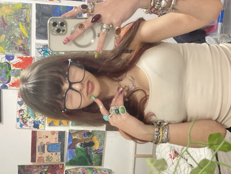 A woman with brown hair, bangs, and glasses takes a selfie in front of an art-covered wall. She wears a cream-colored tank top and displays numerous rings and bracelets. Her nails are painted with colorful designs. She holds an iPhone with a clear case in her left hand, adorned with several rings. A plant is partially visible in the bottom right corner.