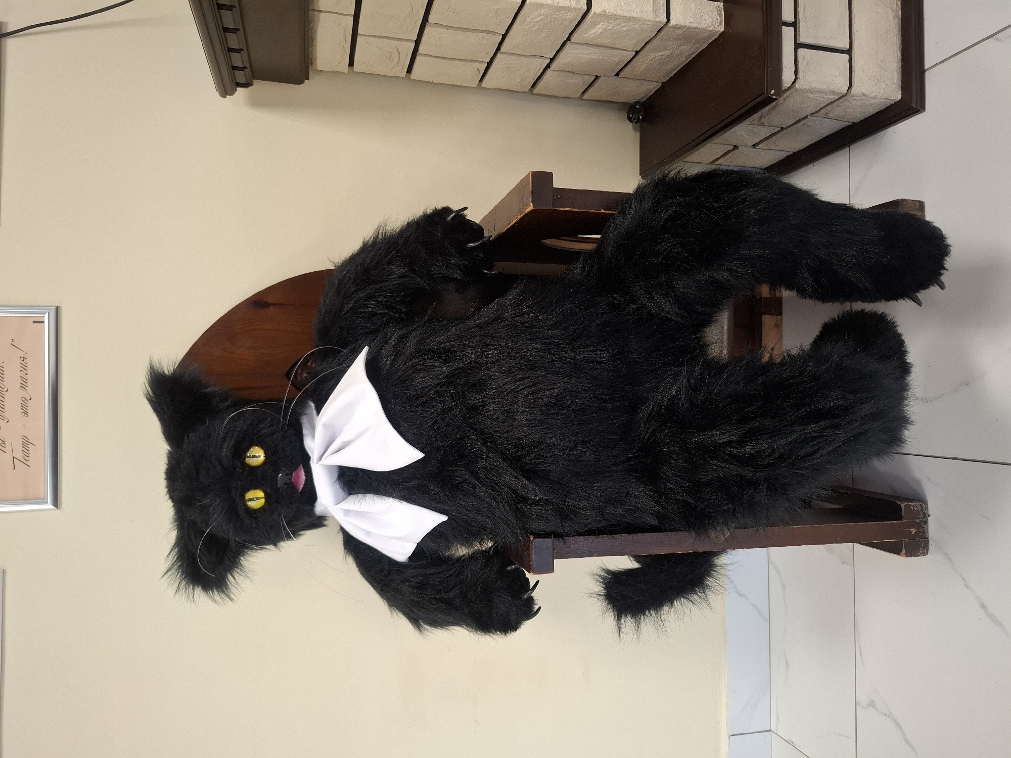 A plush black cat doll with yellow eyes and a white bow sits on a dark wooden chair. The doll has long, shaggy fur and large paws with visible claws. It is positioned in front of a pale yellow wall. To the right of the doll is a decorative fireplace with a brown mantelpiece and a facade made of light-colored bricks. A framed picture hangs on the wall above the doll. The floor is tiled with a white marble pattern. The scene is indoors, likely in a public space given the framed text on the wall, possibly a theater or cultural center in Baykalsk, Russia.