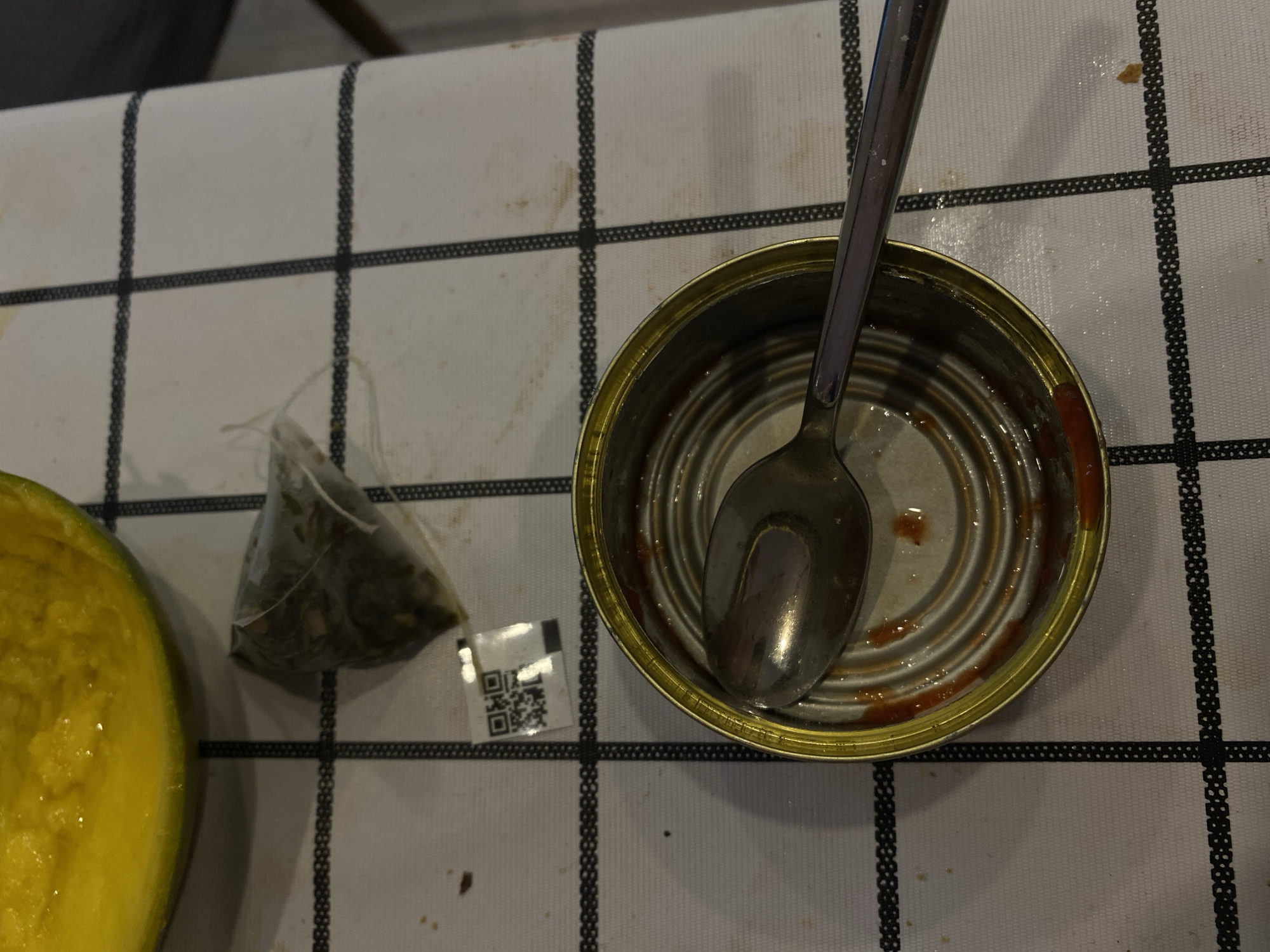 The image shows a close-up, overhead view of a white, checkered tablecloth with some scattered crumbs and smudges. On the tablecloth, there is a slice of a yellow fruit, possibly mango or melon. To the right of the fruit is a small, transparent tea bag filled with dried leaves. Below the tea bag is a small white label with a QR code. A used, silver spoon rests inside an open, empty tin can, which has some reddish residue on its interior. The setting appears to be indoors, likely a dining table, and does not provide any specific clues that would allow for the identification of its location within the city of Lomonosov, Russia.