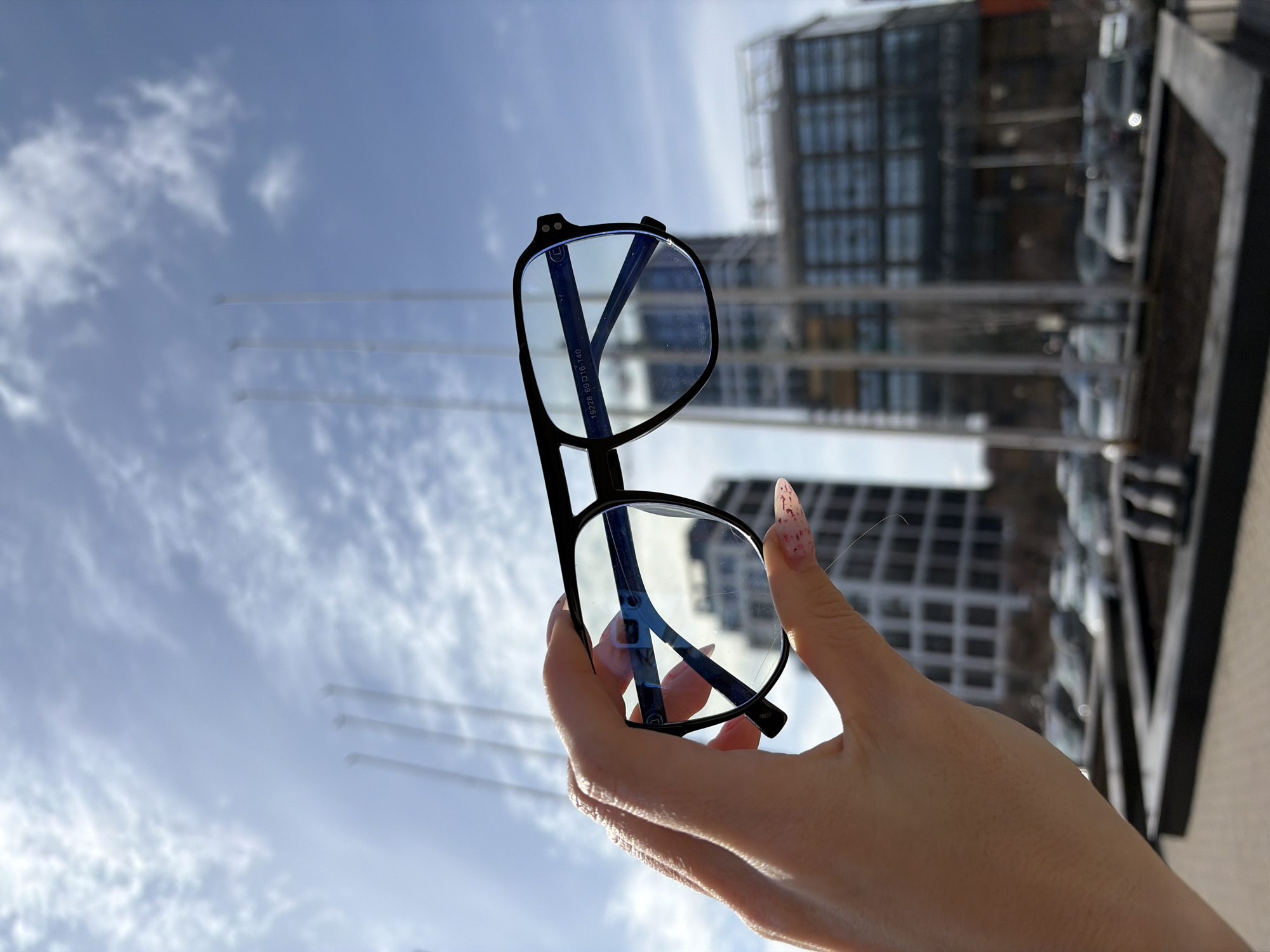 A photograph captures a person holding a pair of black-rimmed glasses against a backdrop of a cloudy sky and an urban landscape. The glasses are the focal point, positioned in the foreground and held up by a hand with long, painted nails. Through the lenses, the background is visible, revealing a modern building with a grid-like facade and several tall, slender poles extending into the sky. In the distance, there is a hint of other buildings and parked vehicles. The shot appears to be taken from a low angle, looking upwards towards the sky and the buildings. The city is Izhevsk, Russia.
