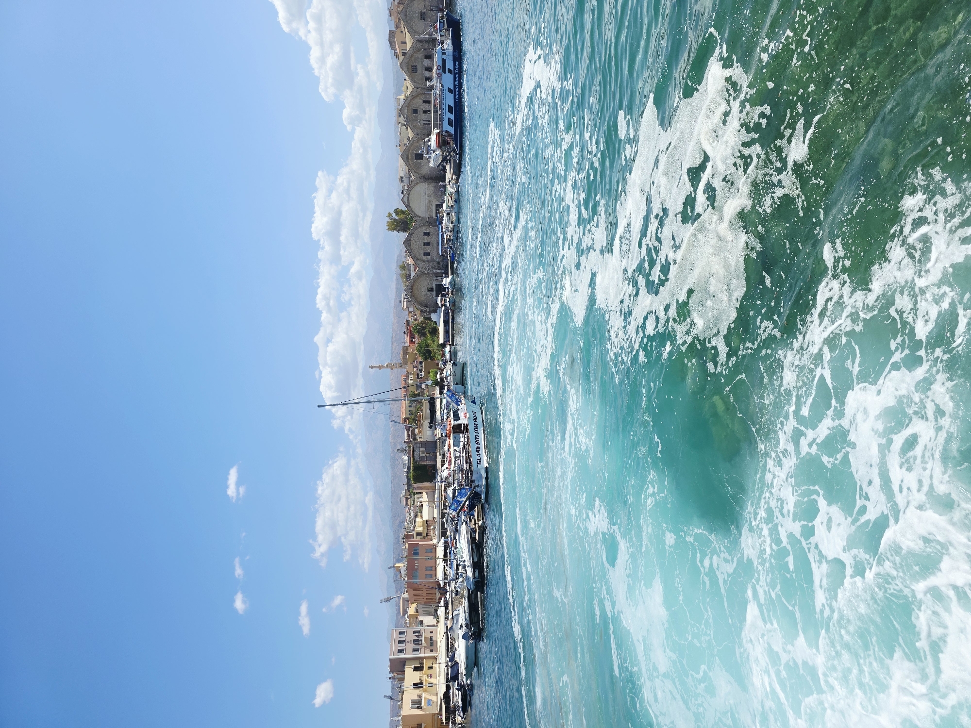 This is a high-angle, eye-level, full, medium shot of a harbor in Chania, Greece, under a bright blue sky with scattered white clouds. In the foreground, turquoise water churns with white foam, indicating the wake of a boat. Several boats are docked in the middle ground, including a white and blue "GLASS BOTTOM BOAT" with "CHANIA BOAT TRIPS" written on its side. Behind the boats, buildings with light yellow and beige facades line the harbor. The architecture suggests a historical port area, with a row of arched, stone buildings in the background. Mountains are visible in the distance. The scene is devoid of people.