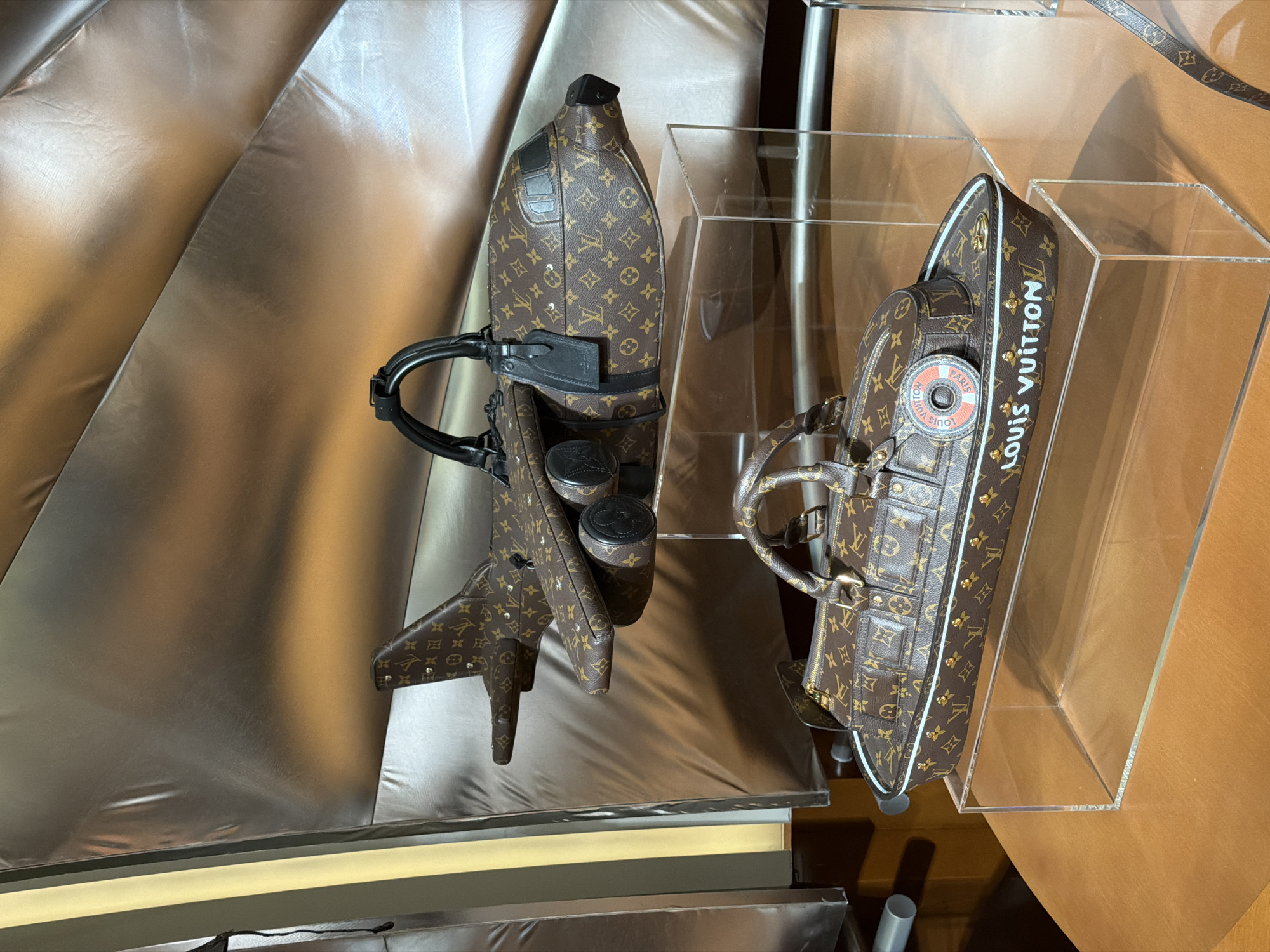 The image depicts two Louis Vuitton bags, each crafted in the shape of a boat. The bags are on display, one positioned on a silver, reflective surface and the other enclosed in a clear acrylic case. The bag outside the case is detailed with black straps and cylindrical accents, while the one in the case features a circular design with "Paris" inscribed. "Louis Vuitton" is written vertically on the case side. The environment suggests an upscale retail setting, possibly within Jing'an District, China, based on the style and product being showcased.