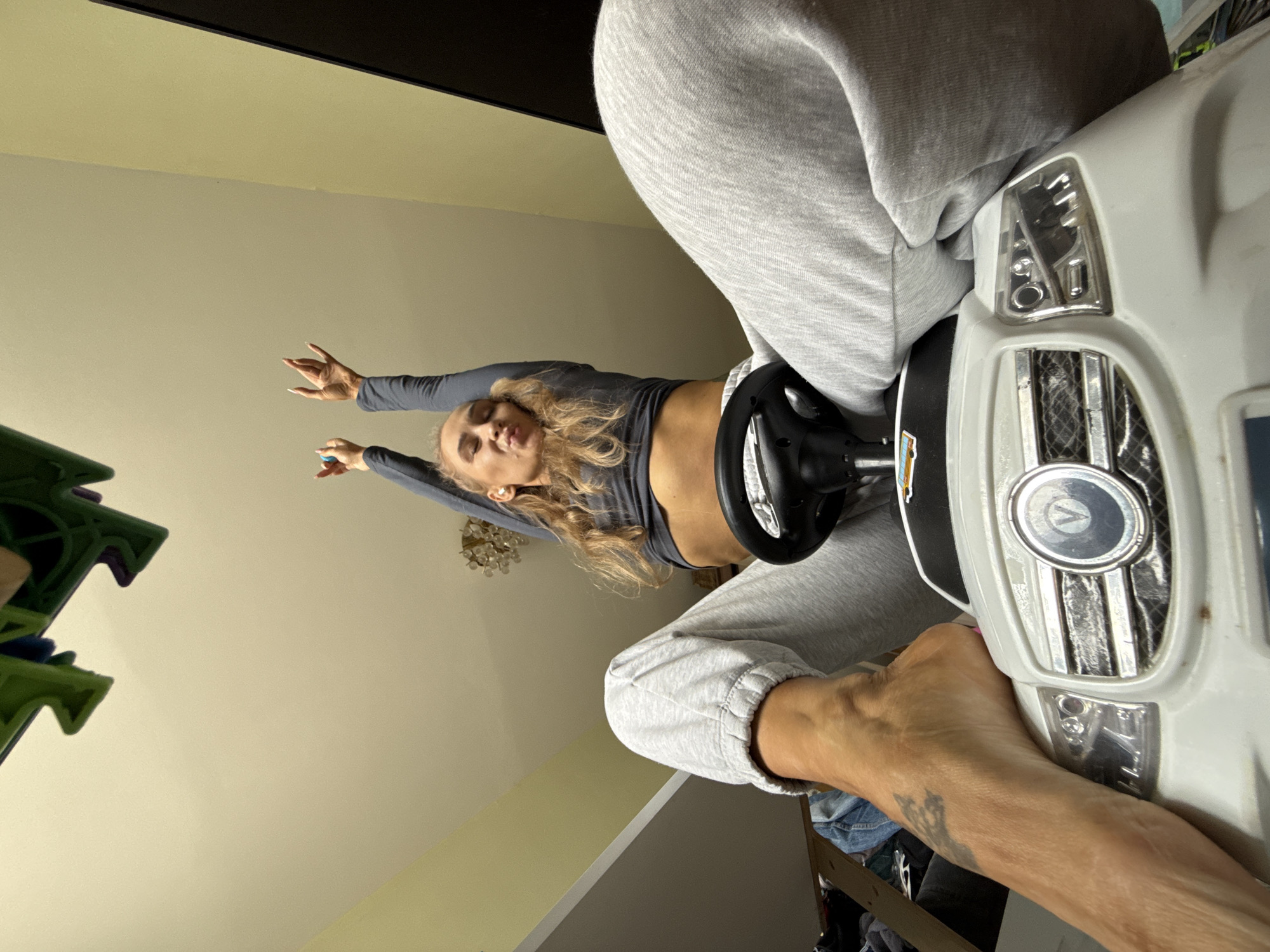 An indoor, low-angle shot captures a young woman engaging playfully with a white child's ride-on toy car. The woman, with long, wavy blonde hair, wears a dark grey long-sleeved crop top that exposes her midriff, and light grey sweatpants. Her torso is arched backward, arms extended outwards with open hands, and her lips are pursed in a playful "pout" expression. Her legs are draped over the toy car, and her bare right foot, adorned with a dark tattoo resembling a crown, rests on the front grille of the car. Her long fingernails are visible.
The toy car features a black steering wheel, clear plastic headlights, and a textured grille. In the background, light-colored walls (beige/cream) are visible, along with a partially obscured green structure on the left and a glimpse of a chandelier or decorative light fixture. A dark, possibly black, screen or frame runs along the top edge of the image. The overall mood conveyed is one of lightheartedness and fun. The scene is set indoors, in what appears to be a casual living space or play area. No visible text is present. The location context is Chaikovsky, Russia.