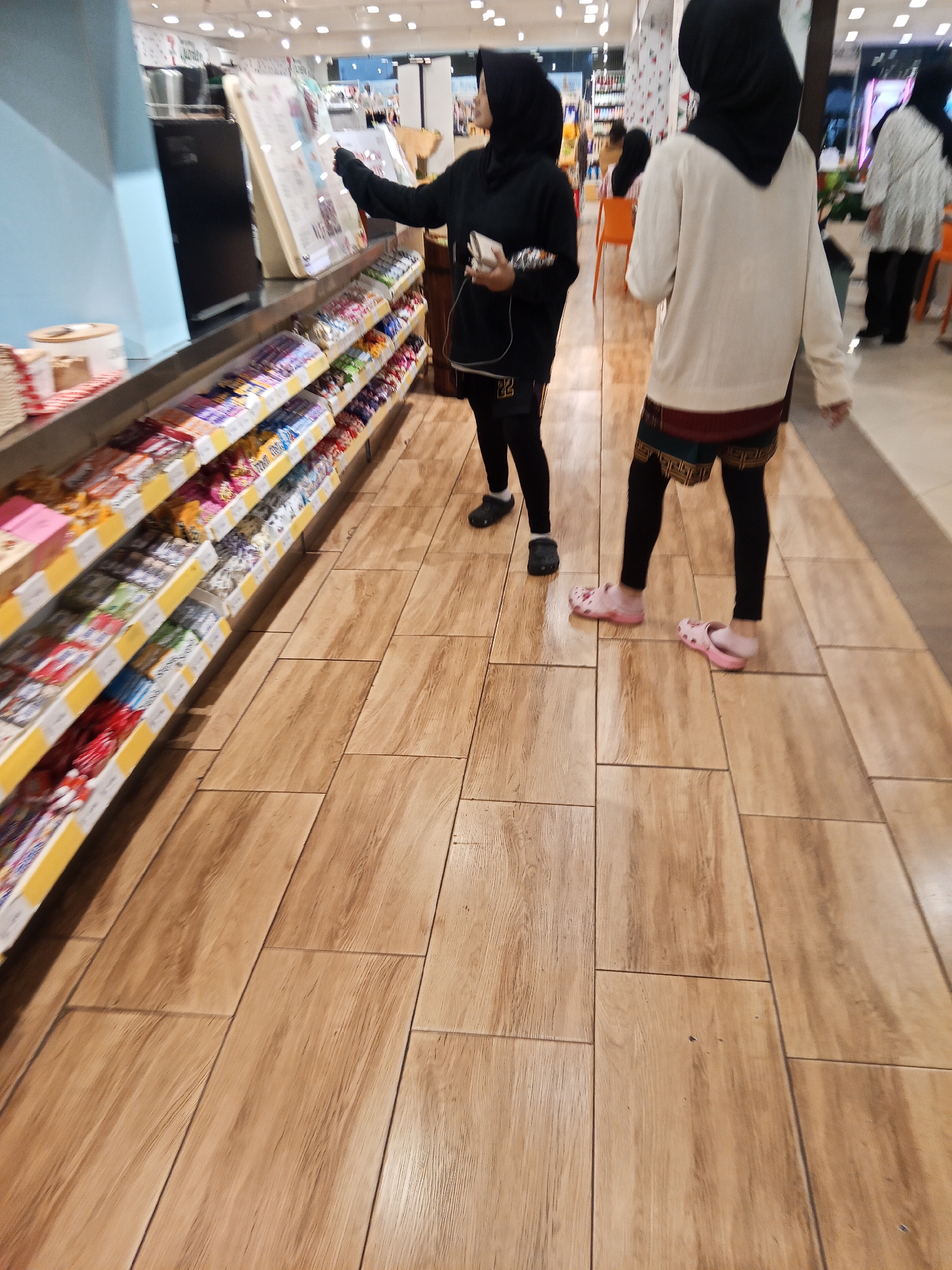 The image shows an interior view of a store, likely a convenience store or supermarket, with aisles of products displayed on shelves. The flooring appears to be wood-look tiles. In the foreground, two individuals wearing headscarves are visible. One, on the left, is wearing a black headscarf and black clothing, holding a small white object. The other, on the right, is wearing a cream-colored top and black leggings with patterned shorts, and pink slip-on shoes. A third person in a headscarf and patterned clothing is partially visible further down the aisle, and another person in a white top and dark pants is in the background on the right. The store appears to be well-lit with overhead lighting. The specific location in Lubuklinggau, Indonesia, is not discernible from the image itself, but the presence of items that appear to be packaged candies or snacks on the shelves suggests a retail environment.