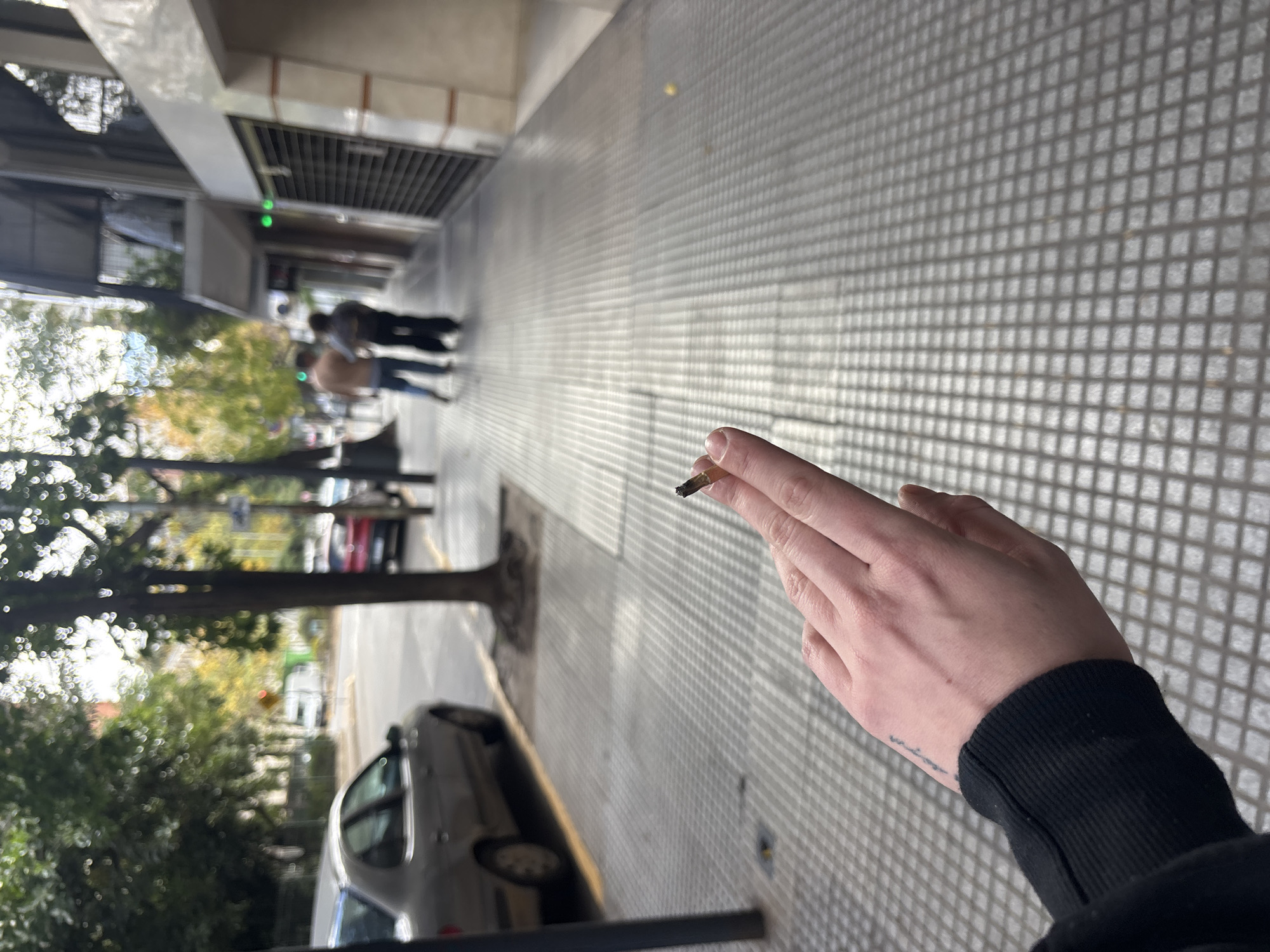 A first-person view from an individual standing on a sidewalk. The sidewalk is paved with small square tiles creating a grid pattern. The individual is holding a lit cigarette between their fingers. The person's left hand is visible, wearing a black long-sleeve shirt. The background shows a street scene with parked cars, trees with autumn foliage, and pedestrians walking in the distance. Buildings line the street, some with storefronts and awnings. The location is in Buenos Aires, Argentina.