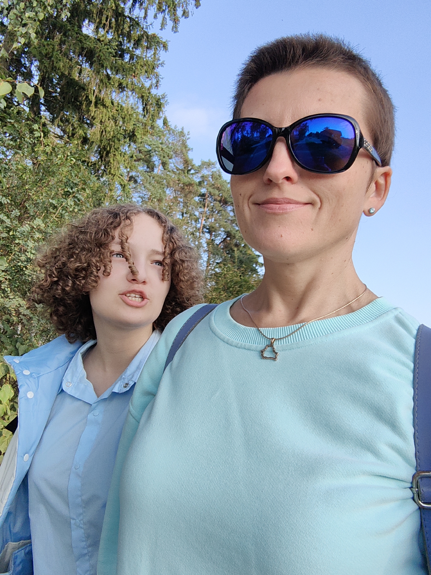 In a close-up selfie, two individuals are pictured outdoors against a backdrop of green trees and a clear blue sky. The person in the foreground, on the right, has short, dark hair and wears large, dark sunglasses with blue lenses that reflect the surroundings. They are also wearing a light blue crewneck sweatshirt with a delicate, irregularly shaped pendant on a thin chain. To their left, a younger person with curly, light brown hair is visible. This individual is wearing a light blue collared shirt, partially covered by a light blue jacket. The setting appears to be a park or wooded area, possibly within the city of Fanipal, Belarus, given the natural scenery. The lighting suggests it is daytime, likely late afternoon due to the warm tones and angle of the light.