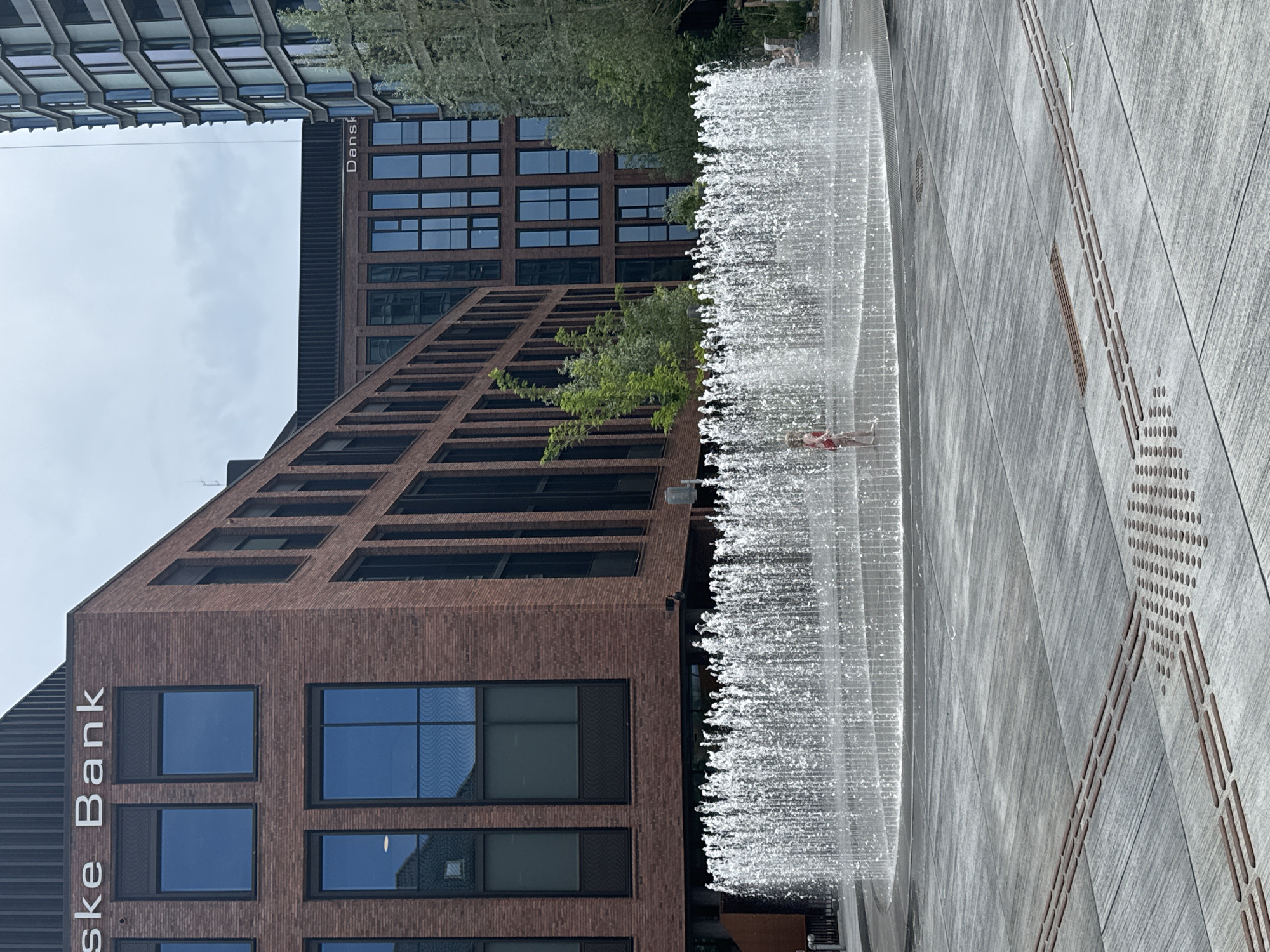The image depicts a public square in Copenhagen, Denmark, likely an urban plaza. In the foreground, a large, circular water fountain is active, with numerous jets of water creating a curtain of mist and spray. The ground surrounding the fountain is paved with large, grey tiles, some of which feature tactile paving for the visually impaired. A child wearing a red swimsuit stands within the fountain, partially obscured by the water jets.
Behind the fountain, a modern building constructed of red brick with dark-framed windows is visible. The building has the text "Iske Bank" prominently displayed on its facade. Further in the background, another building with a dark facade and numerous windows is seen, bearing the text "Danske" on its upper section. To the right, a contemporary tall building with a glass facade rises, and trees with light green foliage are present. The sky above is overcast.