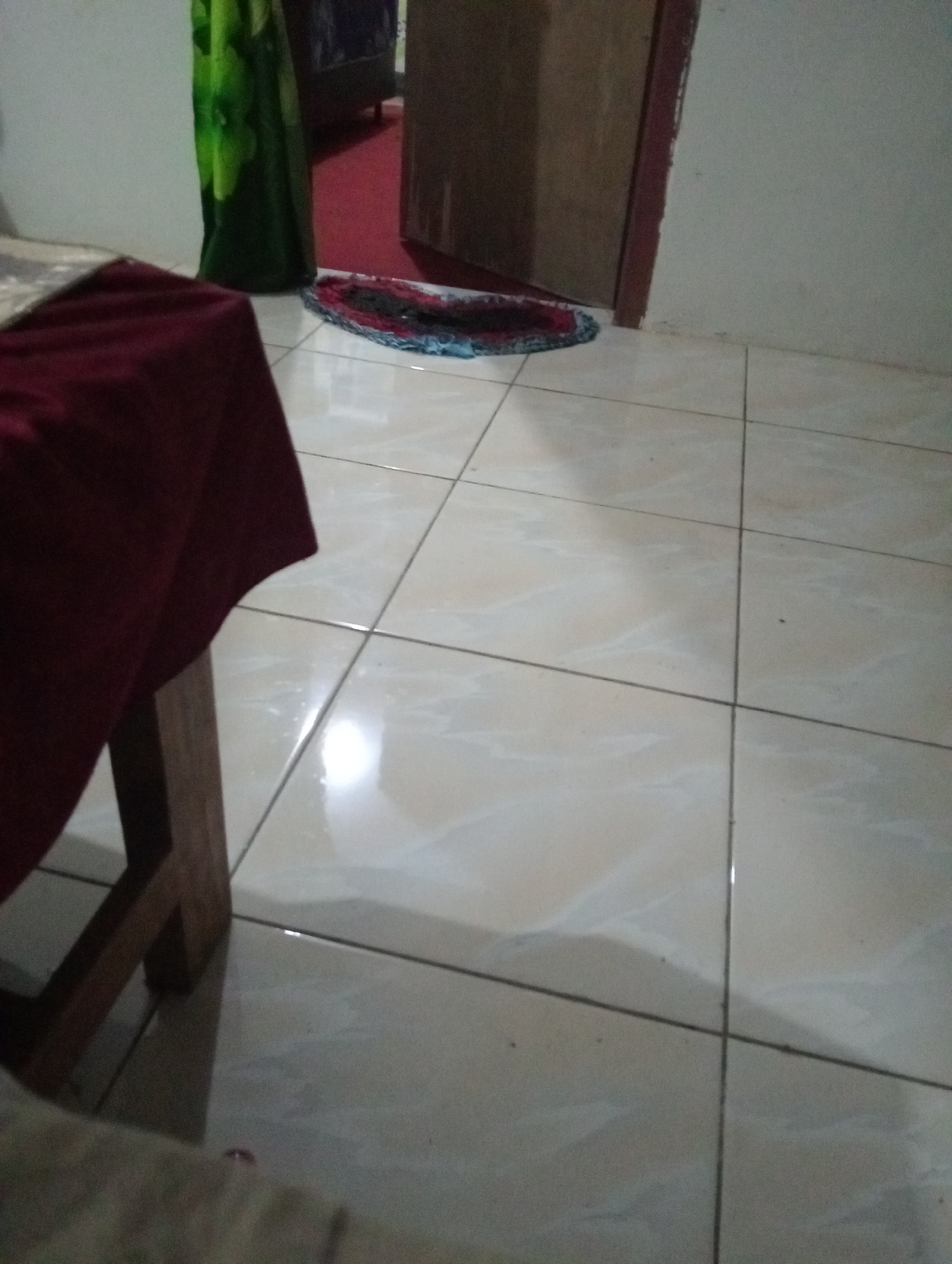 This image depicts an indoor scene, likely a room within a residential property in Sidomulyo, Indonesia. In the foreground, a portion of a table draped with a dark red cloth is visible. To the left, a wooden chair leg is partially visible. The floor is covered with light-colored square tiles with dark grout lines. In the background, a partially open door with a brown frame is visible. To the left of the door, a dark green curtain or fabric is hanging. A small, multi-colored mat is placed on the floor near the door. The lighting suggests ambient indoor illumination.
