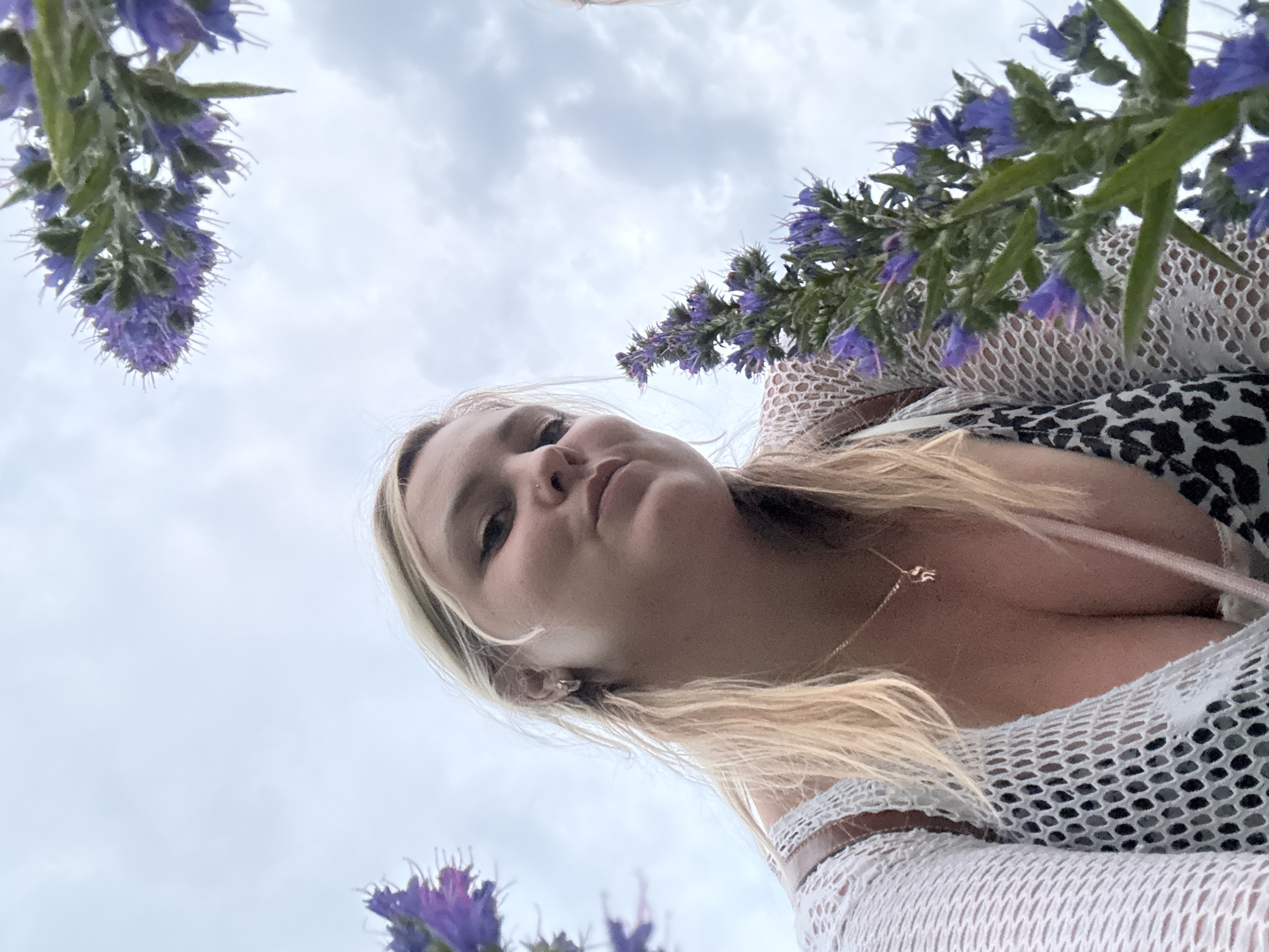 An outdoor, low-angle, close-up selfie shows a woman with blonde hair and light skin, looking up at the camera. She is wearing a white mesh top over a patterned bralette and a delicate gold necklace with a small pendant. Purple flowers frame the shot, with the sky and clouds visible in the background. The image was taken in the city of Chaikovsky, Russia.
