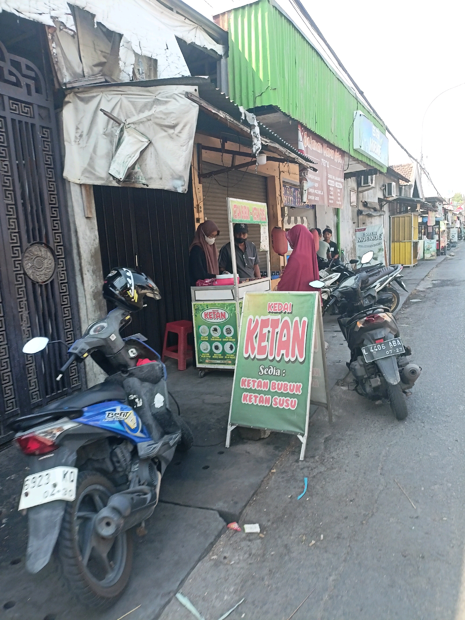 In Surabaya, Indonesia, a street scene depicts a small food stall selling "Ketan" (sticky rice). Two individuals, one wearing a hijab and the other a cap, are visible behind a counter. A sign in front of the stall reads "KEDAI KETAN" and lists "KETAN BUBUK" and "KETAN SUSU" as available items. Several motorcycles are parked on the street, with one blue motorcycle in the foreground showing a license plate with the number "6923 KD". Another motorcycle, dark in color, displays a license plate "L 4406 ABA" with a date "07-27". The stall has a corrugated green roof and is situated on a paved street, with buildings and other stalls lining the road in the background. The overall atmosphere suggests a typical roadside market in the city.