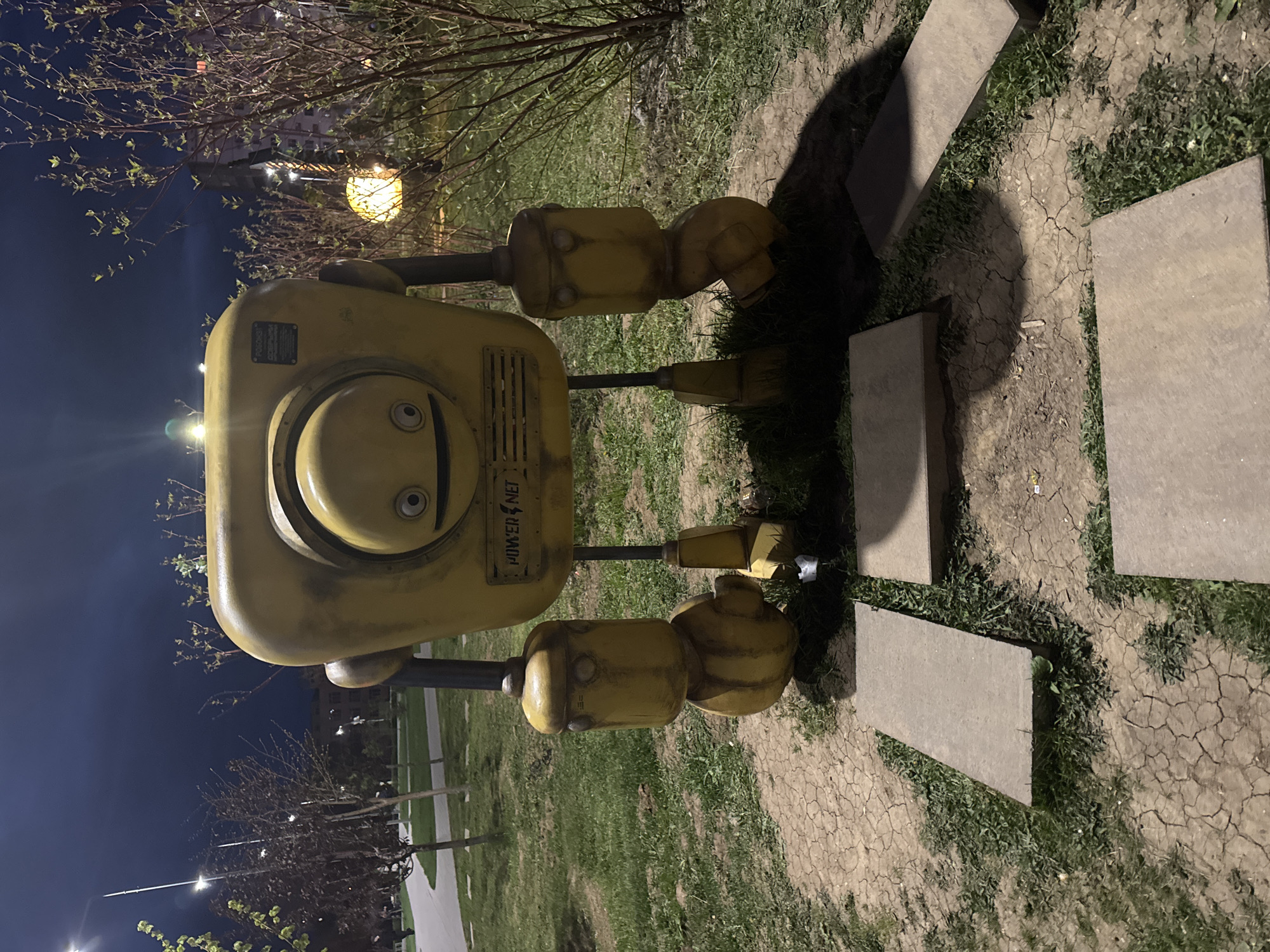The media depicts a large, stylized yellow robot sculpture positioned outdoors in what appears to be a park or public green space at night in Volzhsky, Russia. The robot, which has a somewhat industrial yet friendly appearance, features a rectangular body, cylindrical arms and legs, and a circular "face" with a wide, smiling mouth and two simple round eyes. Its surface shows signs of wear and weathering.
The robot stands on ground covered with sparse green grass and cracked, dry earth. Several rectangular concrete paving stones are laid out around its base, forming part of a pathway. In the background, deciduous trees, some with bare branches and others showing early spring buds, are silhouetted against the dark night sky. Artificial light sources, including bright streetlights in the distance and a glowing spherical lantern nestled within tree branches, illuminate the scene, creating strong shadows on the ground.
Visible text on the robot's chest plate clearly reads "POWER NET." A smaller, dark plaque is also attached to its upper body, but its text is illegible. The scene is static, capturing the sculpture under the ambient glow of night lighting. No people are visible in the image.