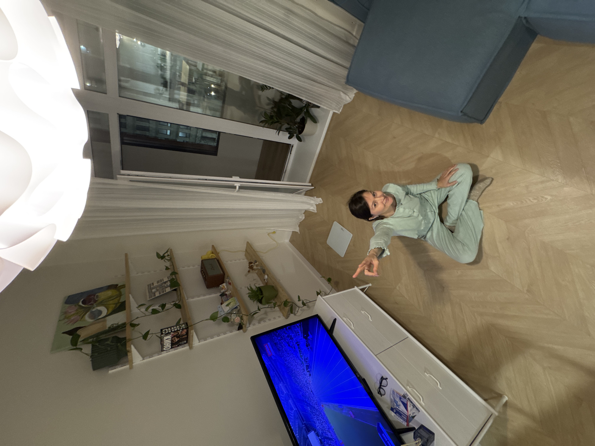 An overhead shot captures an indoor scene, likely a living room, with a woman sitting on a wooden floor. The image is rotated 90 degrees clockwise, giving the impression of viewing the room from above.
The main subject is a woman, wearing a light green, comfortable-looking top and pants, sitting cross-legged on the light-colored wooden floor, which features a herringbone pattern. She is looking upwards towards the camera with a smile and pointing her right index finger, seemingly at the viewer or something out of frame.
The room is furnished with a blue sofa visible in the upper right, and a white dresser or TV stand in the lower right. A large flat-screen TV on the dresser displays a vibrant blue image, possibly from a video game or abstract visuals, with the word "TICKET" faintly visible in the bottom left corner of the screen. Eyeglasses and some papers are also on the dresser next to the TV.
On the left side of the room, near a large window covered by sheer white curtains, a white, sculptural light fixture hangs. Through the window, a dark exterior suggests it is evening or night. A potted plant is placed on the window sill. Below the light fixture, a series of light-colored wooden shelves hold various items including paintings or prints, books/magazines (one with "КИНО" in Cyrillic), a vintage-style radio, and small decorative plants or vines.
The setting appears to be a cozy residential space, likely an apartment in Izhevsk, Russia, given the provided context and the Cyrillic text visible.