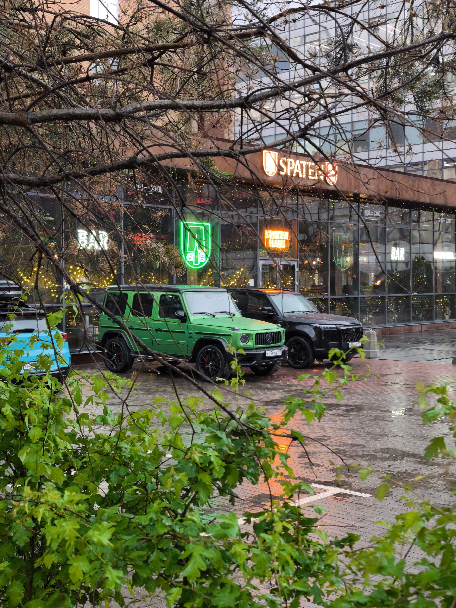 Here is a detailed description of the image:
**Scene:** The photo appears to be taken on a wet, paved street in Moscow, Russia. It is likely early evening, and the street reflects the ambient light and neon signs from a building in the background.
**Environment:** The location appears to be in front of a restaurant or bar called "Spaten Haus" based on the neon signs. The building has a modern glass façade and is adorned with string lights. Wet pavement indicates recent or ongoing rain. Vegetation, including tree branches and bushes, frame the shot in the foreground.
**Objects:**
* Three parked vehicles are visible: a bright green G-Wagon (Mercedes-Benz G-Class), a black Land Rover Defender, and a blue car with a roof rack.
* Neon signs for "Spaten," "Spaten Haus," "G," and "Bar" are visible on the building, indicating the establishment's name and nature.
**Moscow, Russia:** The license plate of the green G-Wagon suggests that it is Russian, specifically from the Moscow region.