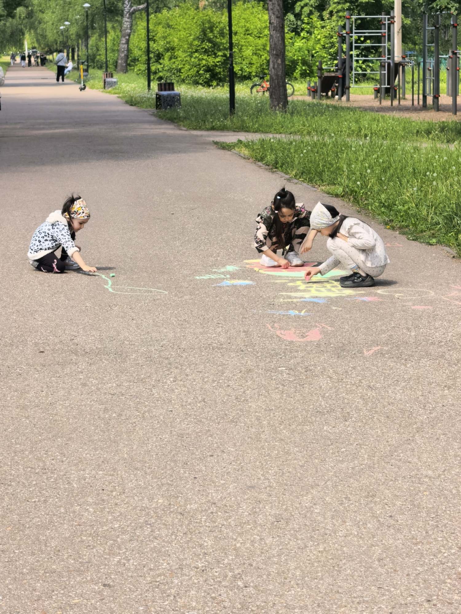 The image shows three young children drawing with colored chalk on a paved path in a park. The child on the left wears a patterned jacket, dark pants, and a headband, and is drawing a green line. The two children in the center and right are squatting and coloring on the path, which already has several colorful chalk drawings. The child in the center wears a patterned outfit, and the child on the right wears a light-colored tracksuit and a head covering. In the background, there is a paved path with people walking, trees, grass, and playground equipment. The location is a park, most likely located in the city of Sterlitamak, Russia.