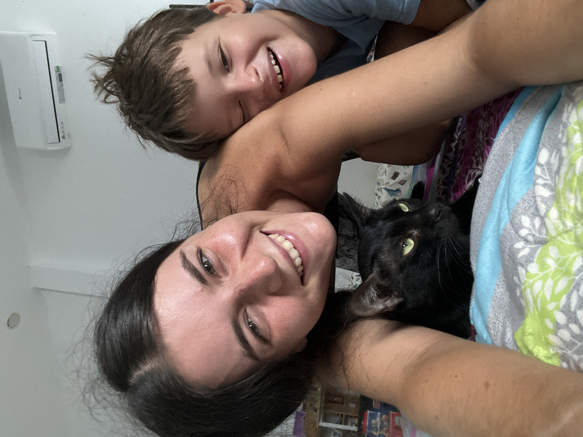 In a bedroom in Mũi Né, Vietnam, a young woman and a young boy are captured in a close-up selfie. The woman, with dark hair and a bright smile, is positioned in the foreground, leaning her head towards the camera. Next to her, the boy is also smiling widely, with one eye winking. Between them, a black cat with striking green eyes is visible, its face partially obscured. The background shows a white wall with an air conditioning unit and a hint of furniture. The subjects are lying on a bed covered with patterned blankets.