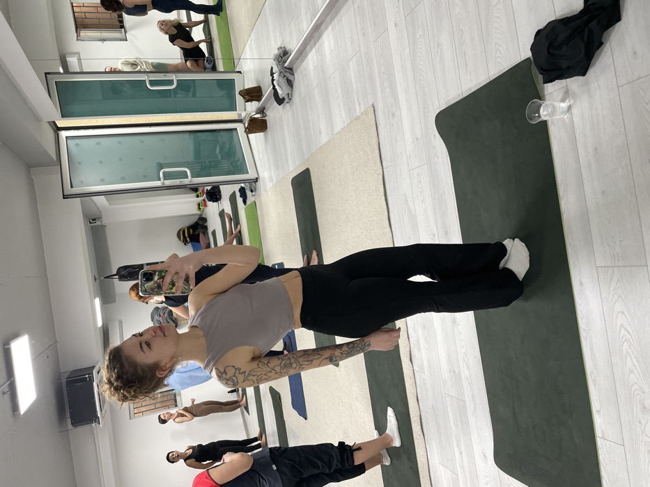 An indoor studio in Budva, Montenegro, features white walls and a light-colored wooden floor. The room is illuminated by overhead rectangular fluorescent lights. A significant portion of the right wall consists of large mirrors, with double glass doors leading to an adjacent area.
The primary subject is a young adult female captured in a selfie, holding a smartphone with a green floral case in her right hand. She has light, curly hair styled in a bun, visible tattoos on her left arm, and is wearing a light grey sleeveless crop top, black flared yoga pants, and white socks. She stands on a dark green yoga mat, looking towards the camera with a slight smile.
In the background, several individuals are present within the studio. To the left of the primary subject, one person in dark clothing and white socks stands on a yoga mat. Further left, another person in dark activewear and one in a light brown jumpsuit are also standing. Reflected in the mirrors on the right, a man in a light-colored sweater is visible through the open glass doors, and two women are inside the studio: one is positioned low on a yoga mat, potentially using a foam roller, and another stands upright.
Multiple dark green yoga mats are laid out across the light wooden floor, interspersed with some beige rugs. A black cylindrical object, possibly a punching bag or fitness equipment, hangs in the background. Personal belongings, including bags and shoes, are scattered along the back wall. In the immediate foreground, next to the primary subject's mat, a clear glass filled with water and a folded black garment are on the floor.