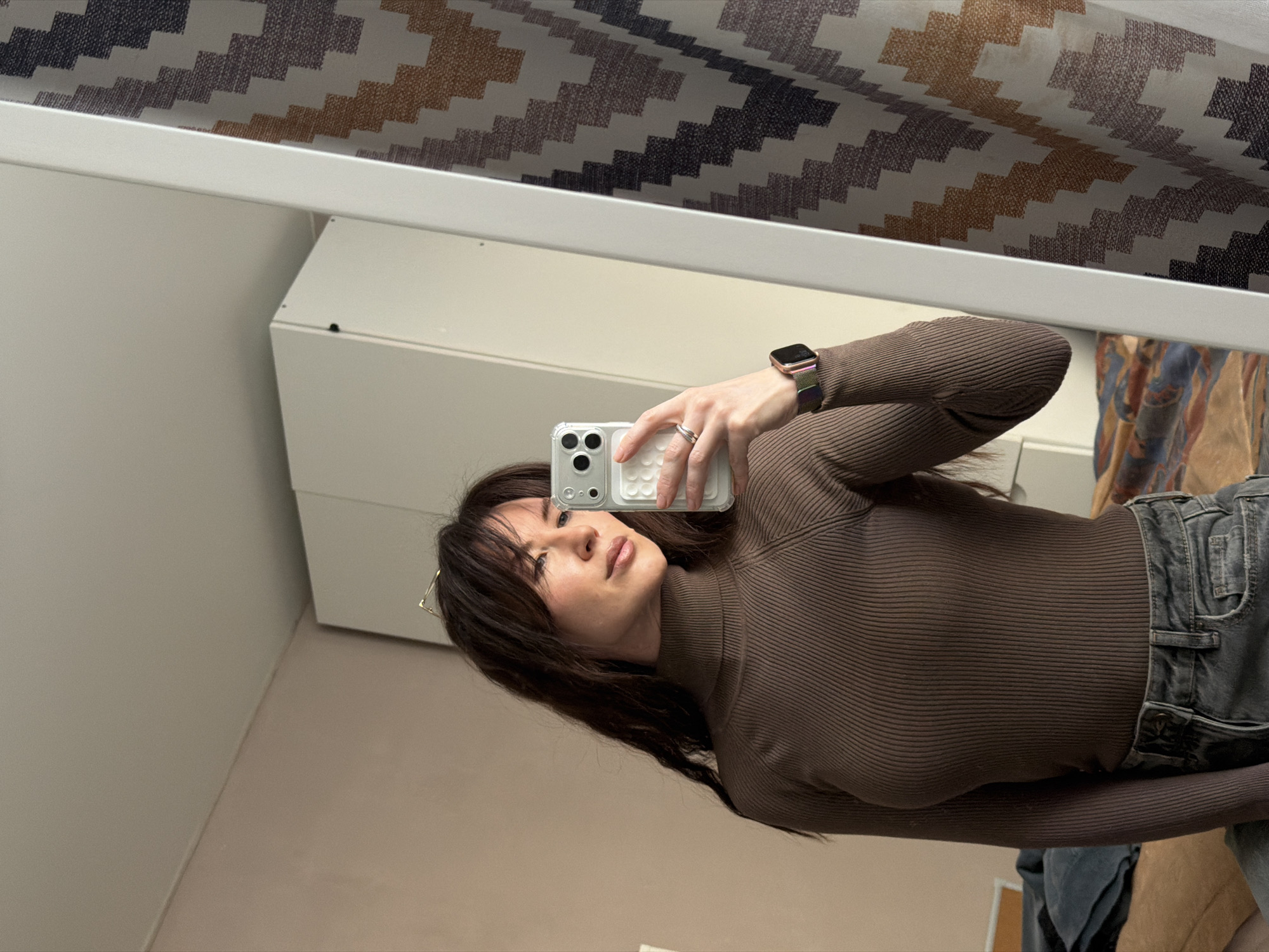 A female subject is depicted taking a mirror selfie indoors in Perm, Russia. She has long, dark brown hair with bangs and is dressed in a form-fitting, ribbed, dark brown turtleneck top tucked into light gray denim jeans. A silver-toned ring is present on her left ring finger, and a smartwatch with a multi-colored fabric band is worn on her left wrist. A pair of gold-rimmed glasses or sunglasses are perched on top of her head. She holds a smartphone, fitted with a light-colored, possibly textured case, in her right hand to capture the image. Her face is turned slightly to her right, away from the direct camera lens, with a neutral expression. The reflected background features a light beige wall and a white ceiling to the left, alongside a white cabinet. To the right, a curtain with a zigzag pattern in brown, orange, purple, and white hues is visible, partially obscured by the white frame of the mirror. Below the subject's waist, a bed covered by a multi-colored patterned blanket and a crumpled dark fabric is discernible.