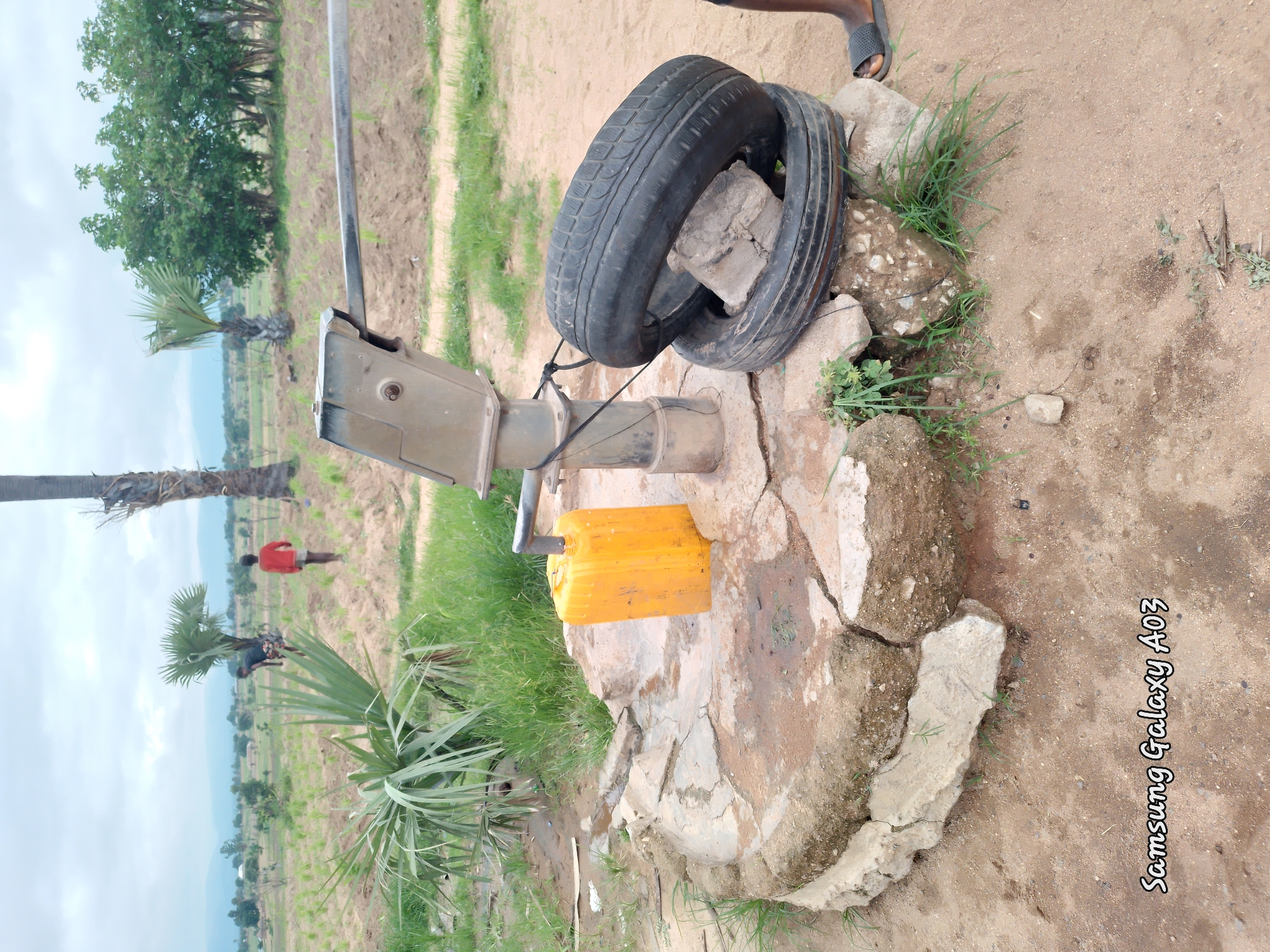 In Angwan Hausawa, Nigeria, a scene unfolds around a manual water pump. The pump, made of weathered metal, stands on a cracked concrete base, its lever arm extended. Adjacent to it, a bright yellow jerrycan rests, likely filled with water. Two black tires are stacked on the ground near the pump, one positioned on top of the other, with a piece of concrete lodged between them. A thin black rope is tied around the pump's support and the upper tire. The environment is dusty and arid, with sparse green grass growing around the base of the pump and tires. In the background, palm trees and other foliage dot a field, suggesting a rural or semi-rural setting. Two figures, possibly children, are visible in the distance, one in red and another in darker clothing, walking across the field. The sky is overcast with clouds. The bottom of the image displays a watermark reading "Samsung Galaxy A03".