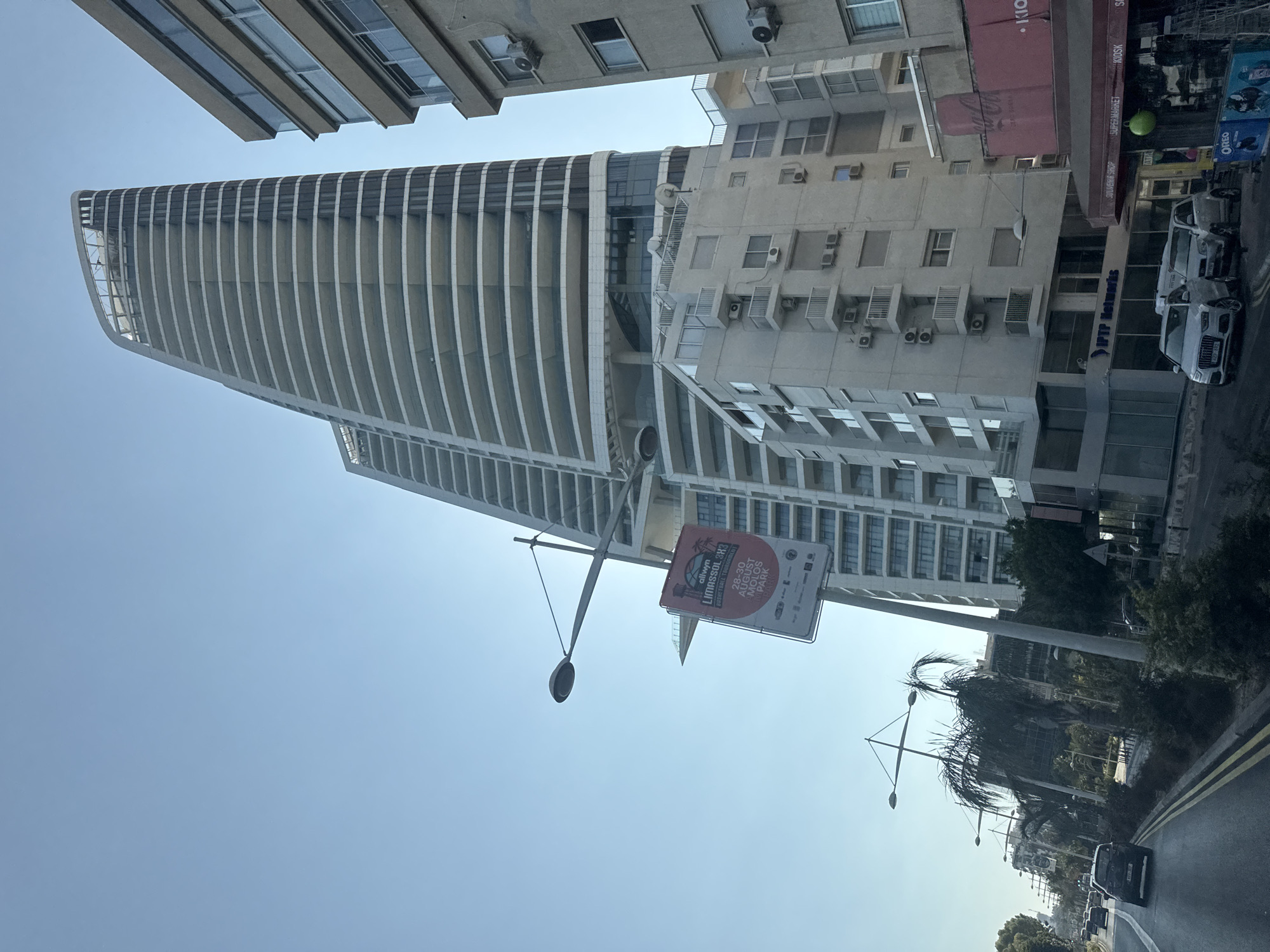 The image captures a street view in Limassol, Cyprus, featuring a distinctive modern skyscraper with a curved facade and numerous balconies. To the right of the skyscraper is a lighter-colored, older building with visible air conditioning units. A banner advertising "Alvwin Limassol 3x3 Basketball Tournament" hangs from a lamppost in the foreground. The street is lined with palm trees and other greenery. Several cars are visible on the road, including a white Audi SUV and a white van. Shops with signage like "SOUVENIR SHOP," "SUPERMARKET," and "KIOSK" are present along the street. The sky is clear and blue.