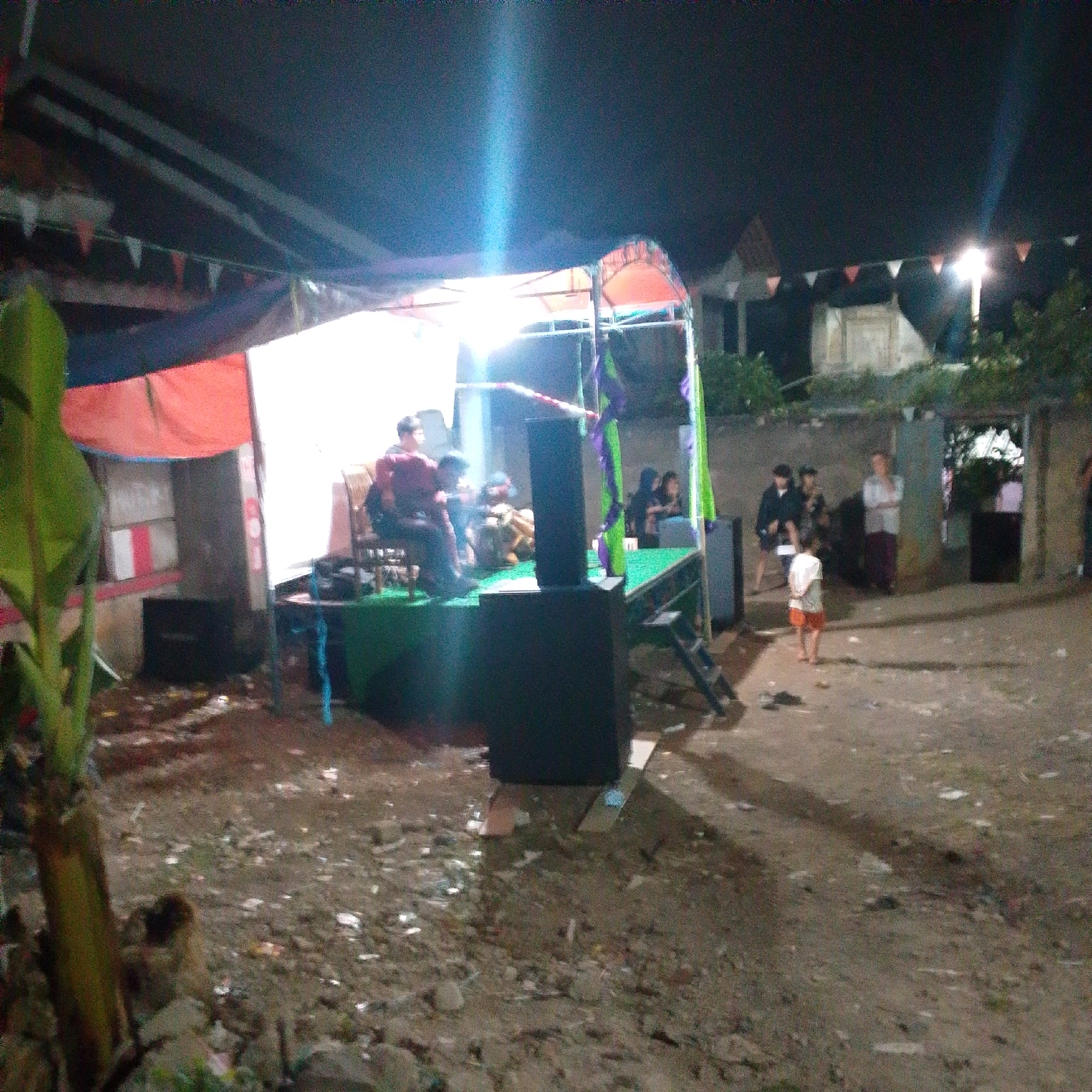 An outdoor, nighttime scene in Cilaku, Indonesia depicts a makeshift stage area. A group of individuals, some seated on chairs and others standing, are gathered on the elevated stage which is covered in green material and adorned with purple and green fabric streamers. A large black speaker is positioned in front of the stage. In the background, a building with a light fixture is visible, and small triangular flags are strung across an overhead structure. The ground is dirt with scattered debris. One person, a child wearing a white shirt and orange shorts, is standing alone in the foreground. Several other people are visible in the mid-ground, some standing and some walking.