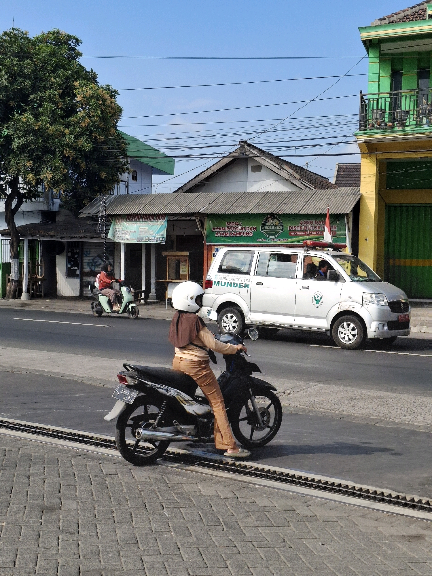 In Lumajang, Indonesia, on a sunny day, a street scene unfolds. The foreground features a person riding a black motorcycle, wearing an orange hijab and matching brown pants, with their right foot resting on the ground. A second person on a light green electric scooter, also wearing a hijab and orange jacket, is to their left, heading in the same direction. Behind them, a silver ambulance with red lights on its roof and the text "AMBULANCE DESA MUNDER" is parked. The ambulance has a logo on its side. The street is lined with buildings, including a structure with a green roof and a bright yellow facade, and another with a grey corrugated metal roof. A large tree with green foliage and some flowering branches stands to the left of the frame. Power lines are strung across the blue sky, and the road surface is asphalt with a patterned brick sidewalk bordering it.