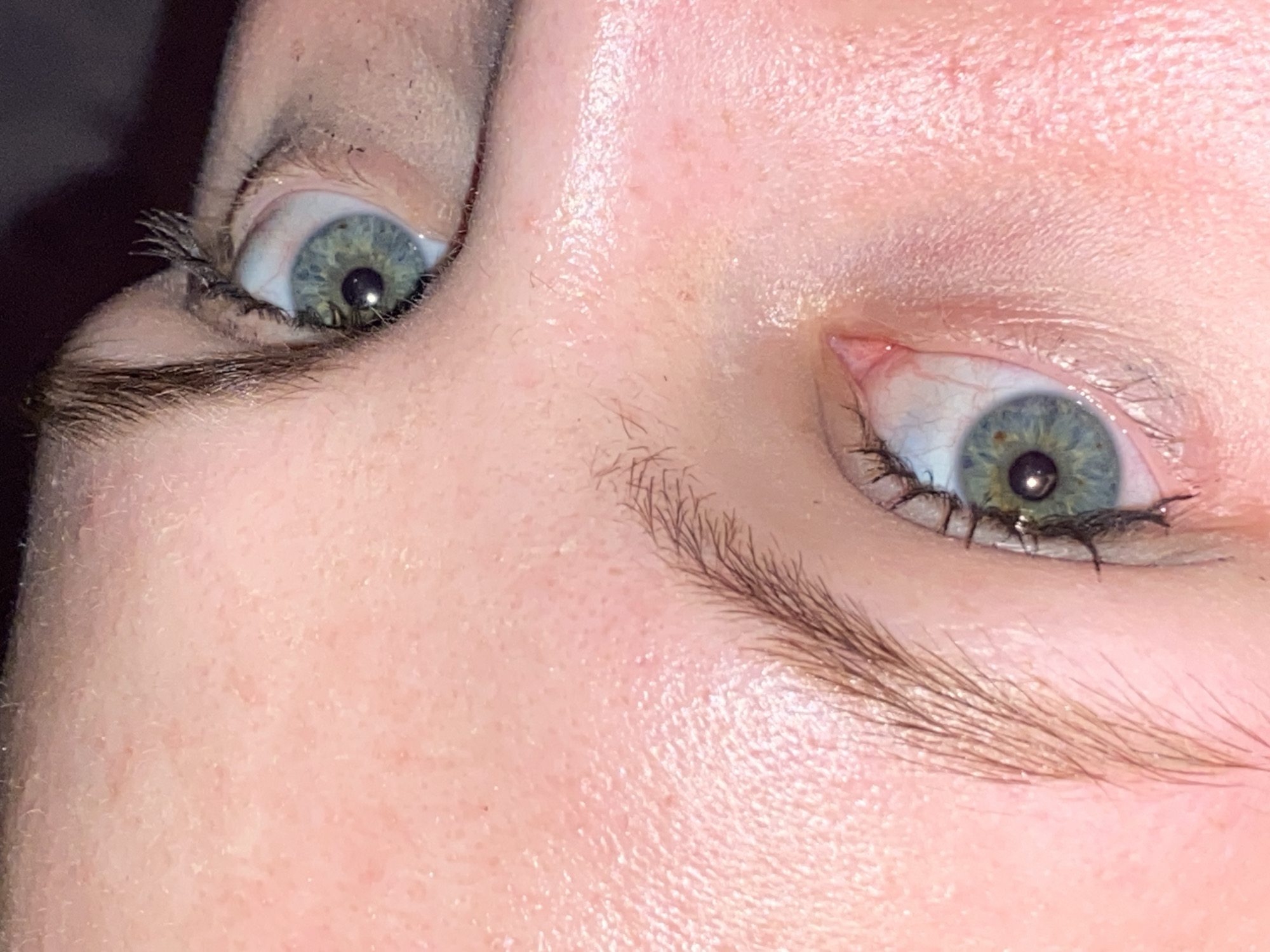 The image is a close-up, low-angle shot of two human eyes, with the subject's face oriented to the right of the frame. The skin tone is fair with visible pores and fine hairs. The left eye, positioned higher in the frame and to the right, is partially visible, showing a blue-green iris with a dark pupil. The right eye, positioned lower in the frame and to the left, is more fully visible, also with a blue-green iris and dark pupil, surrounded by lighter sclera and fine wrinkles. The eyebrows are light brown and appear sparse. The background is dark and out of focus. The image does not provide any discernible information about a specific location in Izhevsk, Russia, nor does it depict any identifiable urban or environmental features.