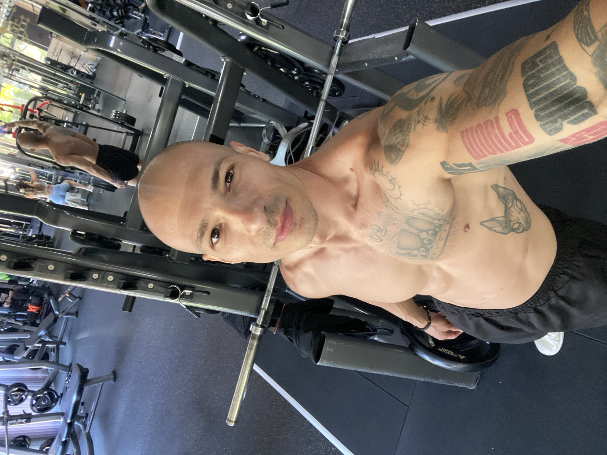 A man, bald with tattoos on his arms and chest, takes a selfie in a gym. He is wearing black shorts and white sneakers. In the background, other people are visible working out, and gym equipment such as weight benches and barbells are present. The environment appears to be an indoor gym with grey flooring and metallic gym apparatus. The image does not contain information that would allow for its location in the city of Pleven, Bulgaria.