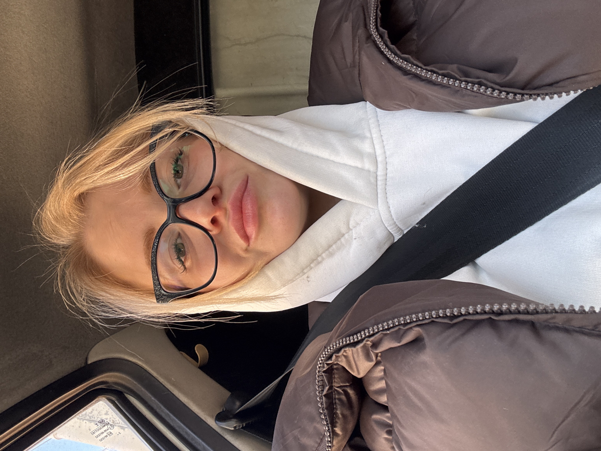 A young woman with blonde hair, wearing eyeglasses with decorative temples, a white hooded sweatshirt, and a brown puffer jacket, is seated in what appears to be a vehicle. A black seatbelt is fastened across her torso. Part of the vehicle's interior, including a window and a section of the dashboard, is visible. The location cannot be determined to be in Saint Petersburg, Russia, based on the provided image.