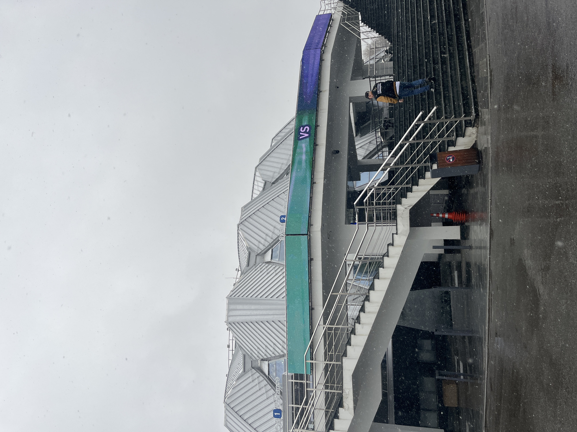 The image shows a modern architectural structure with a striking design, likely a sports venue or arena, situated in Moscow, Russia. The building features a facade composed of intersecting geometric planes covered in a metal siding. A large vertical LED display is affixed to the side of the building, partially obscured by the building structure. The display appears to show a match up with the letters "VS" visible on the screen.
A staircase with metal railings leads to an upper level. A man is visible on the stairs, facing away from the camera, wearing a jacket. The ground is wet, and snow is falling, suggesting a cold, possibly wintry, environment. A traffic cone is on the pavement, presumably for safety. The sky is overcast.