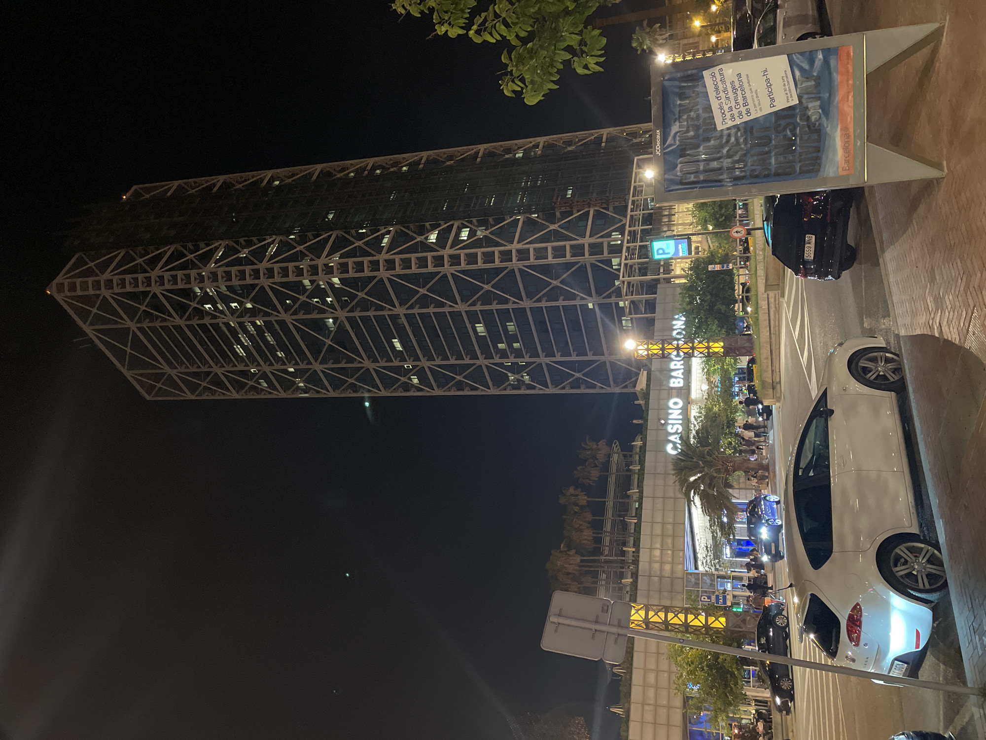 A night-time, low-angle shot captures the Torre Glòries, a distinctive skyscraper in Barcelona, Spain, with its prominent lattice facade illuminated. In the foreground, a white hatchback is parked on the right side of the street, its headlights on. Adjacent to it, a black SUV is visible. Across the street, the Casino Barcelona is illuminated, with palm trees in front of it. Pedestrians are seen walking near the casino entrance. A blue sign with a parking symbol and the word "Taxi" is mounted on a pole. A large billboard with text in Catalan and a picture of a mouth is positioned in the lower right corner of the frame. The sky is dark, with a few faint lights or celestial bodies visible.