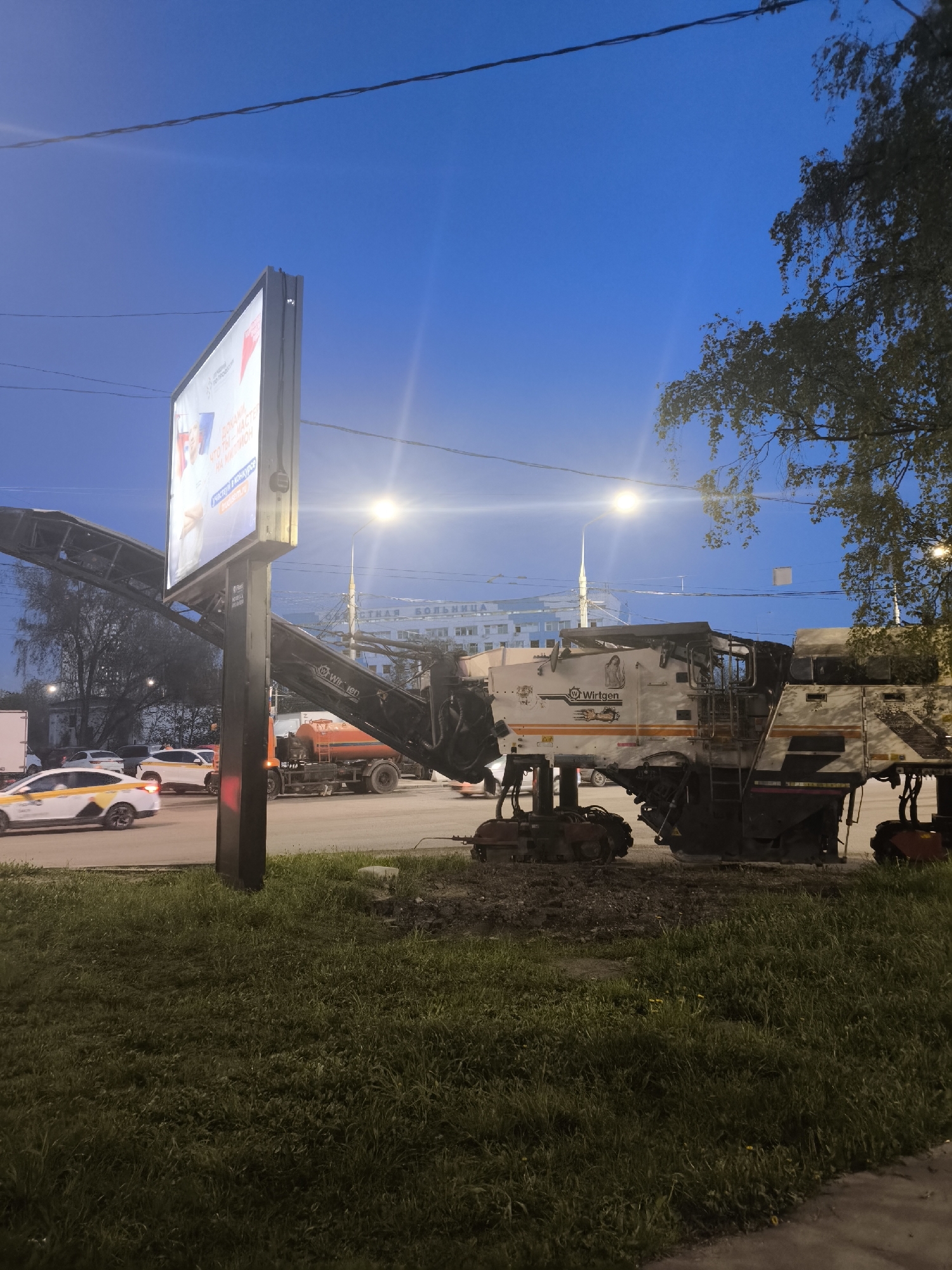 The image depicts a street scene in Khimki, Russia at dusk. A Wirtgen asphalt milling machine occupies the right side of the frame, positioned on a grassy verge adjacent to a road. In the background, several vehicles are visible on the roadway, including white and yellow taxi cabs. A sign post stands on the left, with an advertisement displayed on it. In the far background, a building with multiple spires and a sign reading "Tnaya Bolnitsa" is visible. The sky is a deep blue color, with several street lights illuminating the scene. Overhead power lines are visible.