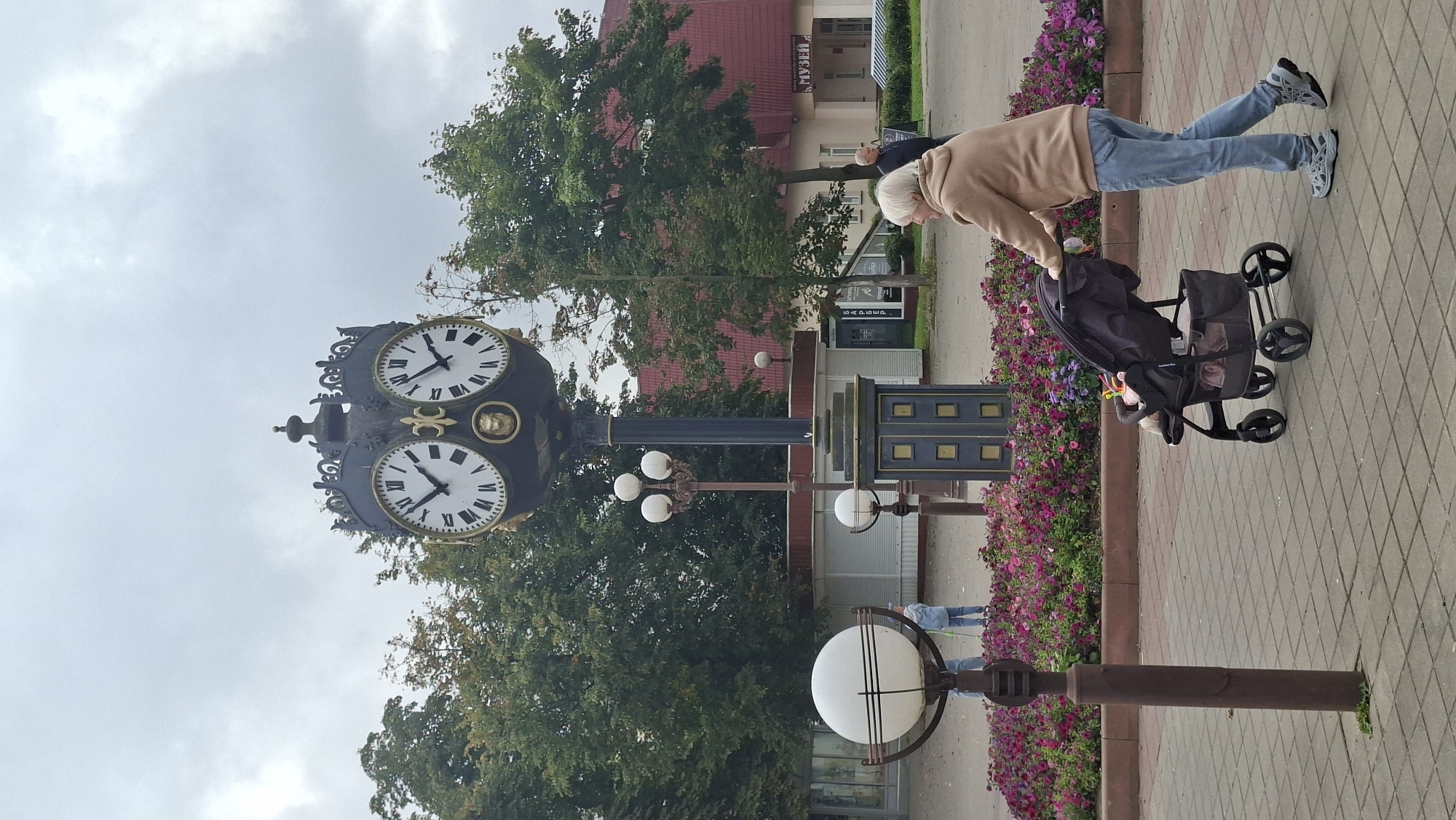 This is a vertical, eye-level, medium shot photograph taken on an overcast day in Maladzyechna, Belarus. The focal point is a large, ornate, double-faced clock tower located in a paved public square. The clock tower is dark blue with gold decorative elements and Roman numerals. To the left of the clock tower is a tall, brown lamppost with three white globe lights. In the foreground, a woman with graying blonde hair is pushing a black stroller with a baby inside. She is wearing a tan hooded sweatshirt and blue jeans. The stroller is positioned on the right side of the frame, angled towards the left. Behind the stroller and to the left of the clock tower is a flower bed filled with pink and purple flowers. In the background, there is a building with a red roof and the word "МУЗЕЙ" (Museum) visible on its facade. Several trees are present, and other people are visible in the distance, including a man walking near the museum and a person with a blue broom near a white kiosk. The ground is paved with square gray and beige tiles.