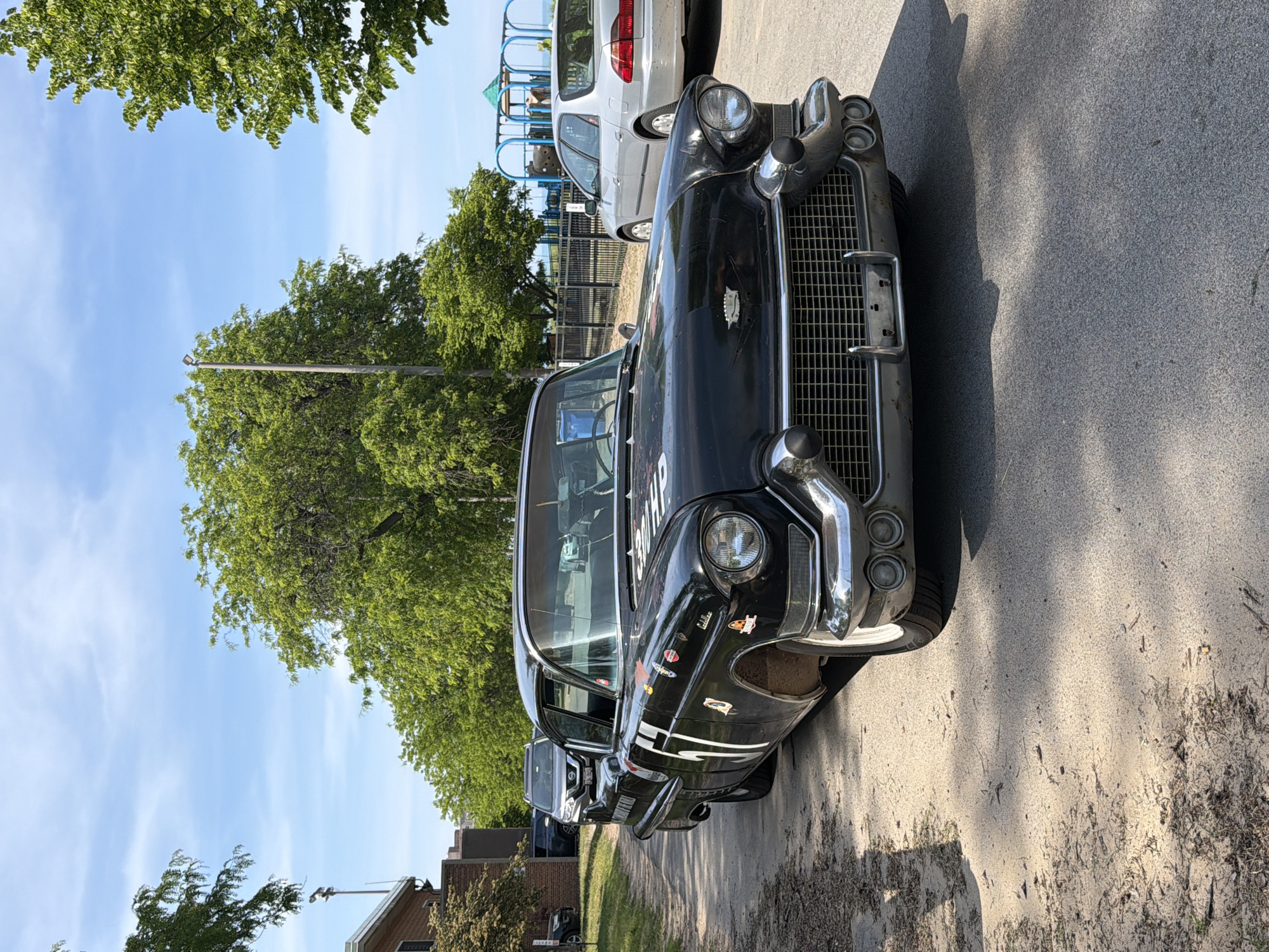 The image shows a black vintage car parked on a paved surface, likely a street or parking lot. The car has chrome accents and is adorned with various stickers. Behind the car, there are other vehicles, trees, and a playground. The sky is blue with scattered clouds. The location appears to be an urban or suburban area within the city of Chicago.