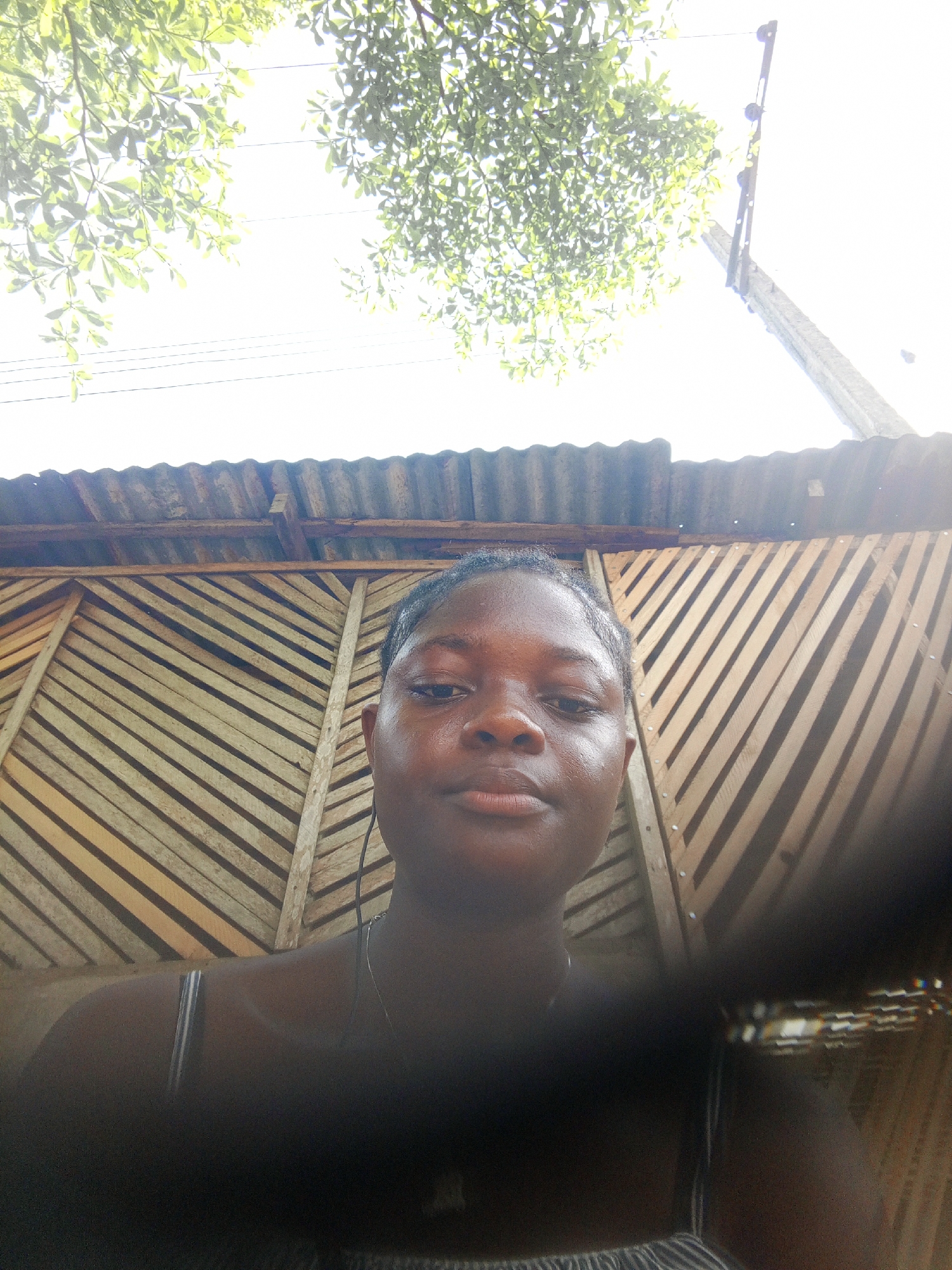 A young, dark-skinned woman is centered in a close-up selfie. She has short hair, a neutral expression, and is wearing a thin necklace. Behind her is a wall made of diagonally placed wooden planks and corrugated metal roofing. Above the wall, there are leafy green trees and a utility pole, likely for power. The setting appears to be outdoors, possibly a semi-enclosed space in the city of Mbribit, Nigeria. The lower part of the frame is partially obscured by a blurred object, potentially the photographer's hand or part of their clothing.