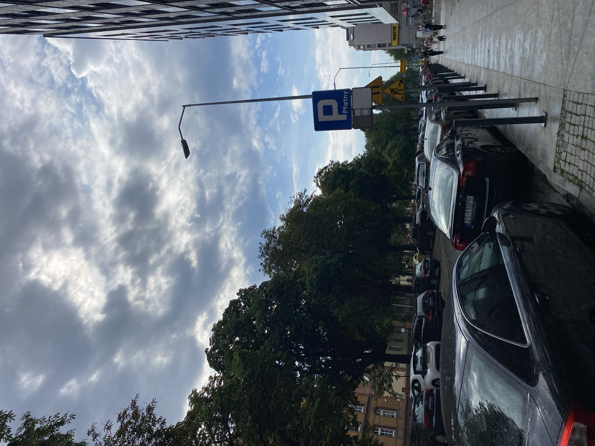 This image captures a street scene in Szczecin, Poland, likely in the downtown area, featuring a row of parked cars along the side of the road. The sky is filled with dramatic, dark clouds, suggesting an impending storm or overcast weather, with patches of blue sky visible and the sun breaking through in some areas.
On the right side of the image, a modern, multi-story building with a dark, reflective facade dominates the skyline. A lamppost stands on the sidewalk, and a blue parking sign with the letter "P" and the word "Płatny" (Polish for "paid") is visible, indicating paid parking. Below it, there are additional traffic signs, including a yellow triangular sign with a construction worker icon.
The sidewalk is paved with concrete and a section of cobblestones, and it is lined with metal bollards. A long line of cars is parked along the street, mostly sedans and SUVs, with license plates visible on some. The cars are of various colors, including black, white, and silver.
On the left side of the street, a row of trees with dense green foliage lines the sidewalk, partially obscuring older buildings with yellow facades and white-trimmed windows.
In the distance, a few people are walking on the sidewalk, and a dog on a leash is visible. The wet appearance of the pavement suggests recent rain or ongoing dampness. The overall impression is of a busy urban street with a mix of modern and older architecture, under a dynamic sky.