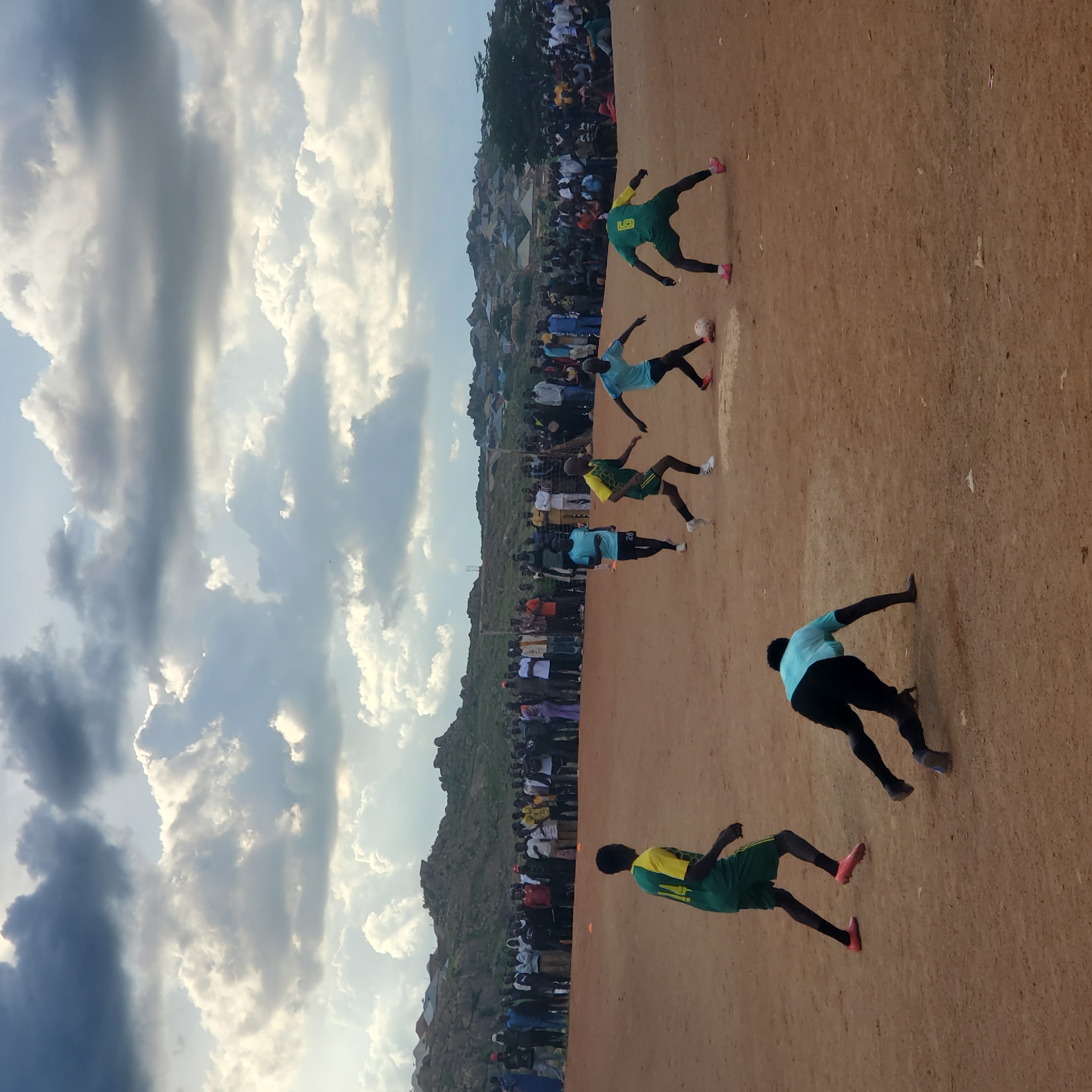 In Jos, Nigeria, a soccer game is taking place on a dusty, outdoor field. The sky above is dramatic, filled with heavy, grey clouds interspersed with patches of bright sky. A large crowd of spectators lines the edge of the field, some standing and others sitting. The field itself is dirt, with a makeshift goal visible in the background.
On the field, several young men are actively playing soccer. One player in a green and yellow jersey with the number 14 on the back is running towards the right side of the frame. Another player, wearing a light blue jersey and black shorts, is in the foreground, on his hands and knees, possibly reaching for the ball which is near him. Further into the field, a player in a green and yellow jersey with the number 6 on the back is dribbling a soccer ball. Another player in a light blue jersey is running towards the left, with another player in a green and yellow jersey in pursuit. The players are wearing soccer cleats. In the distance, a settlement with houses and rocky hills can be seen.