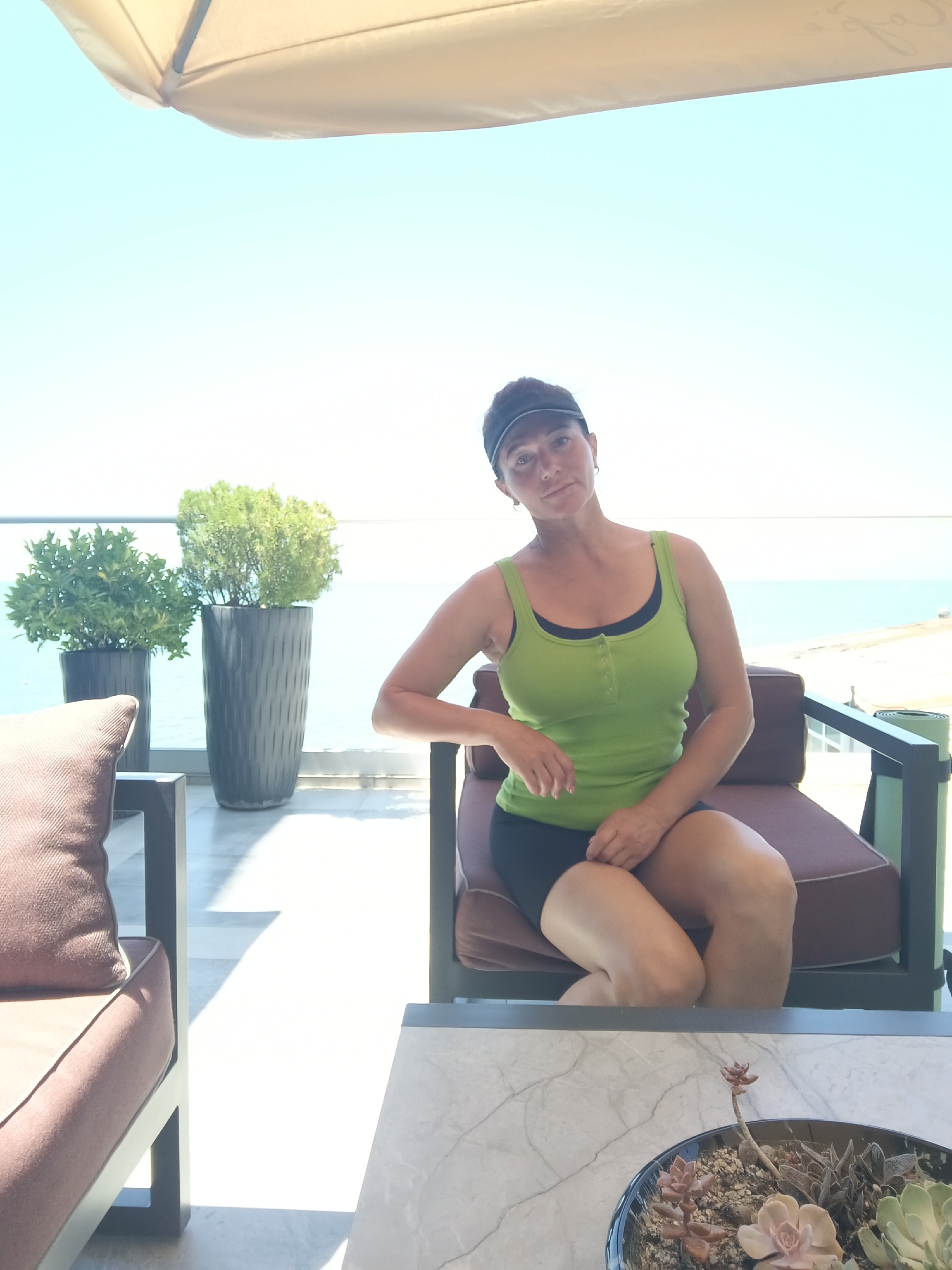 A woman is seated on a chair on a balcony with a view of the sea. She is wearing a green tank top and black shorts, and a black visor. The balcony is furnished with chairs and a table, and there are plants in pots. The scene appears to be set in Sochi, Russia, based on the visual cues of a sunny day and a coastal view.
