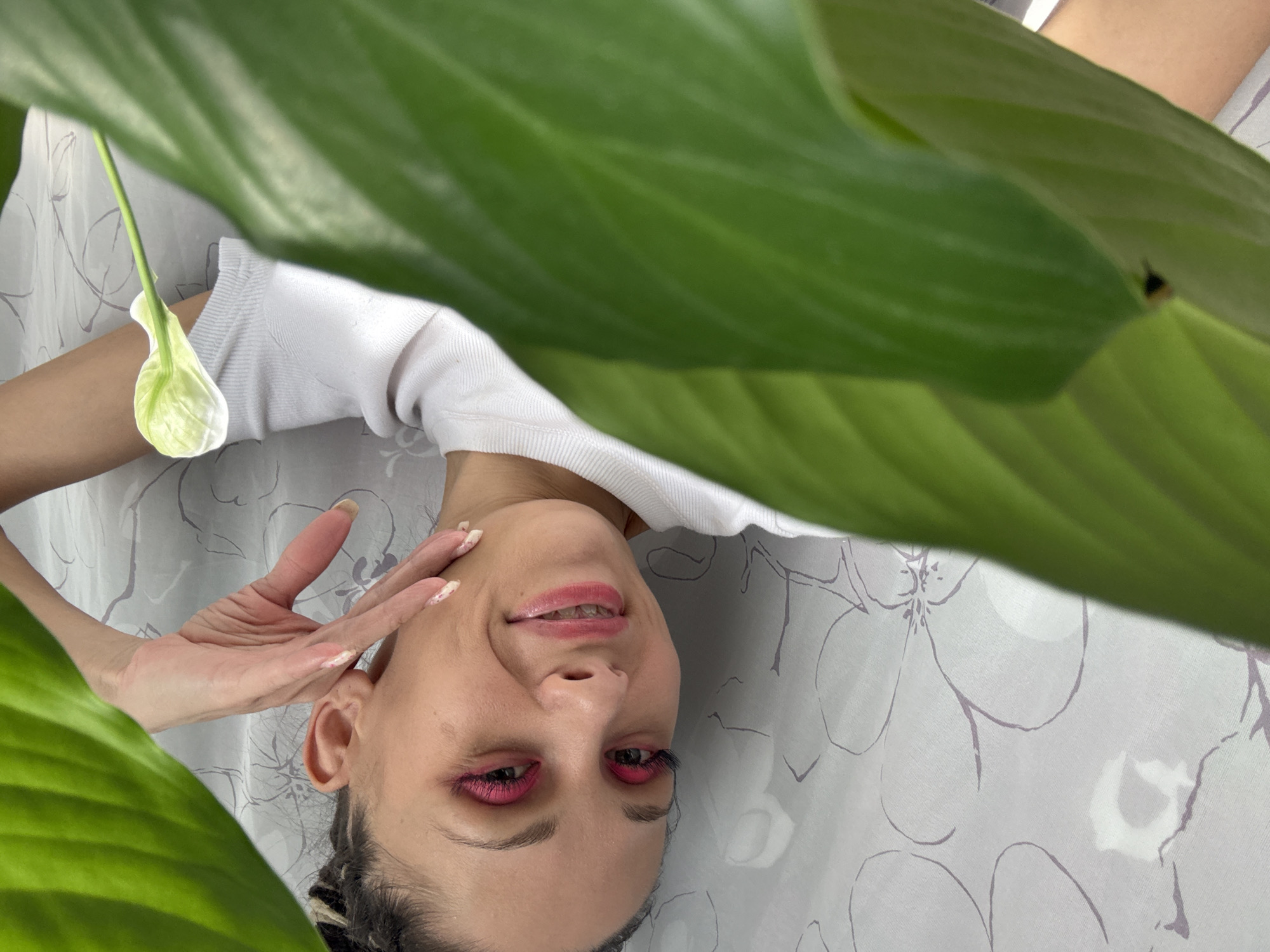 A woman is lying on a light gray floral patterned surface. Her face is visible, positioned centrally in the frame and slightly turned. She has light skin, pink eye makeup, and dark hair pulled back from her face. Her right hand is raised near her face, fingers extended. A white flower hangs near her left shoulder. Large green leaves surround her, partially obscuring her and the background. The location is Rusanovo, Russia.