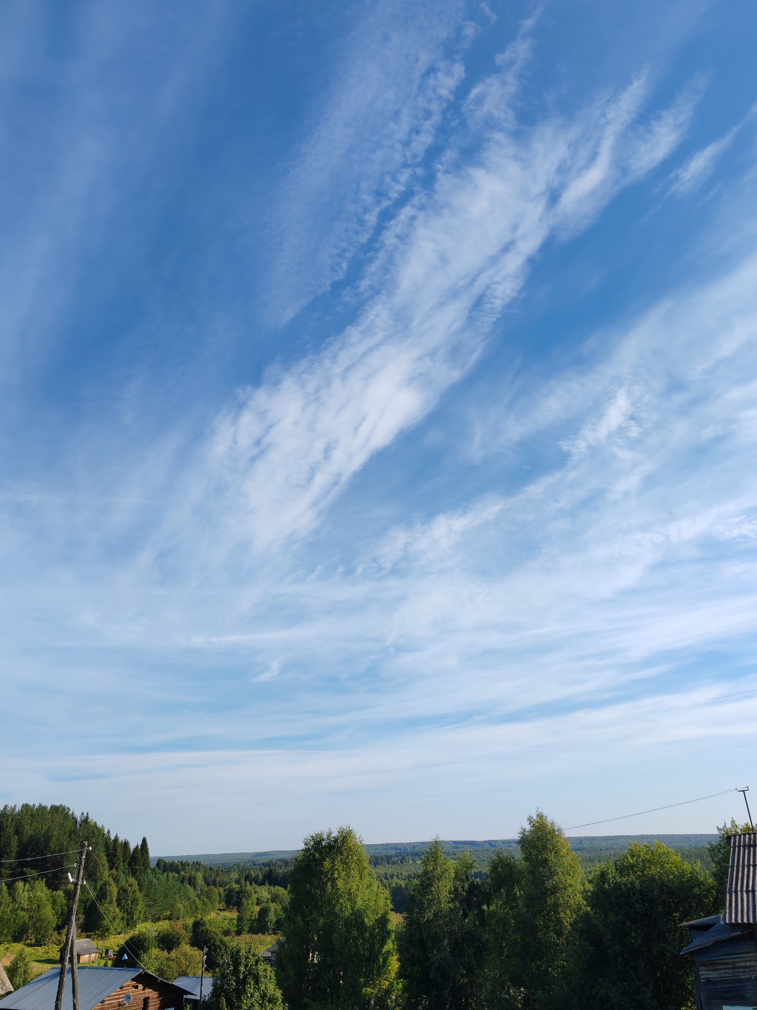 The image captures a rural landscape under a bright blue sky with scattered cirrus clouds. The foreground shows parts of a wooden building with a dark, possibly metal, roof and another structure that appears to be a small shed or outbuilding. A wooden utility pole with wires is visible to the left. Beyond these structures, a dense forest covers rolling hills, with varying shades of green indicating different types of trees and vegetation. The horizon is marked by a further expanse of trees, suggesting a vast, forested area. The overall scene is one of natural beauty and rural tranquility, likely in a location like Чухлэм, Russia, given the typical architecture and abundant forests found in such regions. There are no people or animals visible in the frame, and no actions or interactions are taking place.