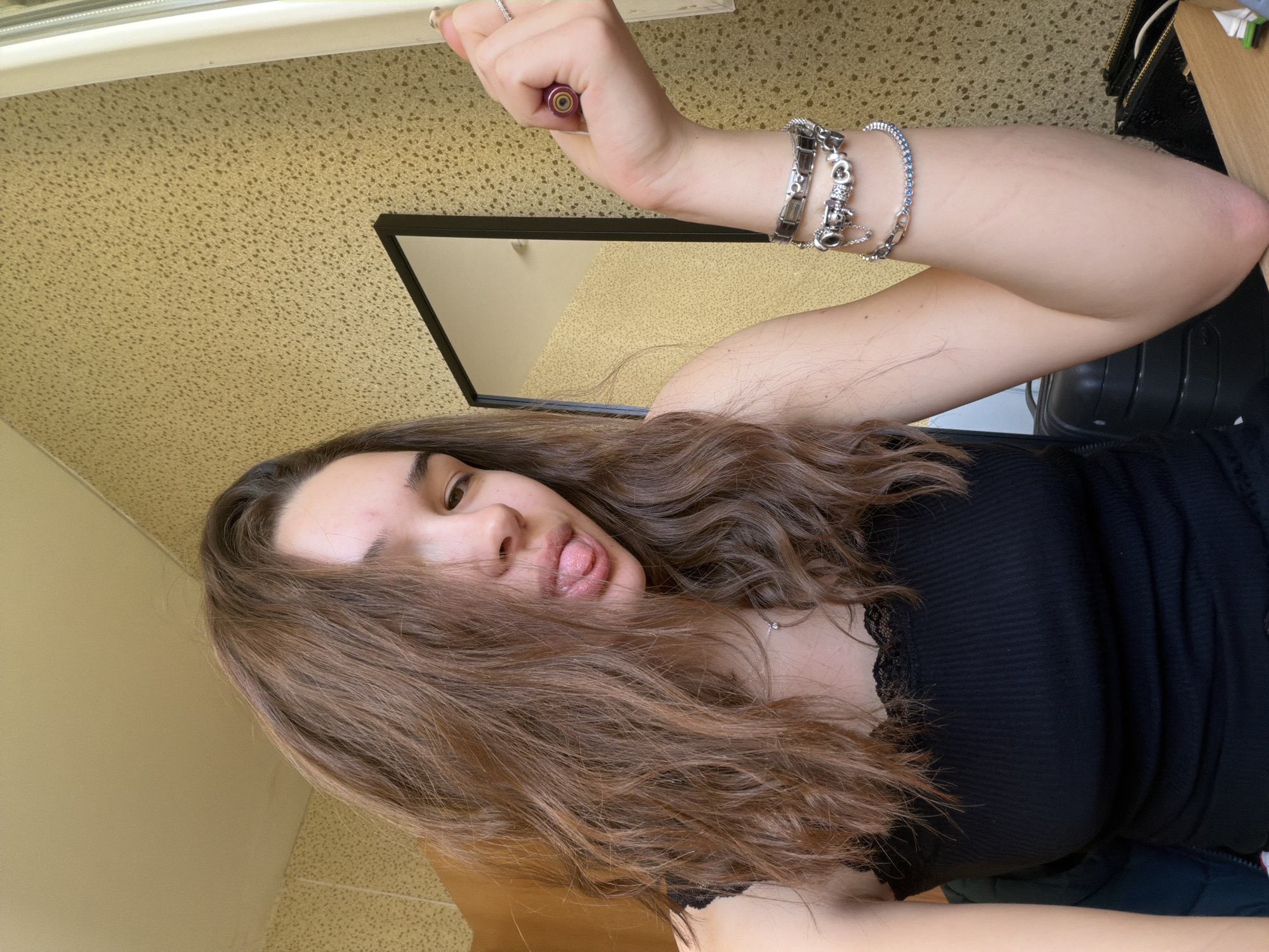 This is an informal, sideways portrait of a young woman, likely a selfie, taken indoors in Sofia, Bulgaria. The woman is positioned with her head tilted, looking towards the viewer with a playful expression, sticking out her tongue. Her long, wavy brown hair falls loosely around her face and shoulders.
She is wearing a black ribbed top with delicate lace trim along the neckline. On her right arm, which is raised, she wears several silver-toned bracelets, including a charm bracelet and one adorned with small blue stones. A ring is visible on her right hand. She holds a small, tubular object with a reddish-purple base and a metallic top, resembling a lip product or vape pen. Faint, linear marks are discernible on the inner aspect of her upper right arm. A delicate silver pendant necklace rests against her chest.
The background features a textured, light brown or beige surface, possibly a wall or ceiling. A mirror with a black frame is visible behind her, reflecting part of the room. Other elements include a dark object, possibly a bag or chair, and a light-colored wooden surface. The lighting is bright, suggesting daytime conditions indoors. There is no visible text in the image. The overall impression is one of casual youthfulness and lightheartedness in a personal indoor setting.