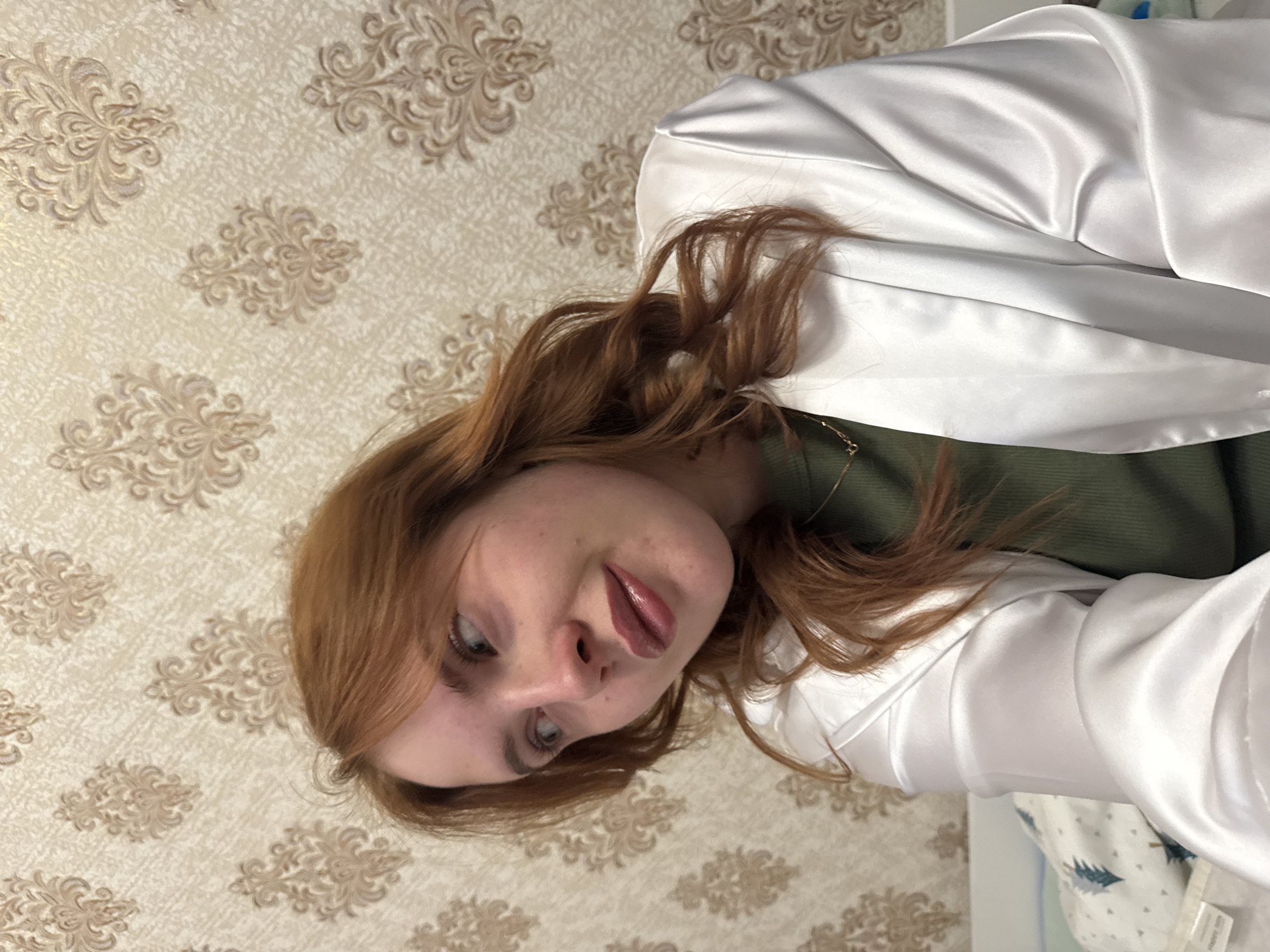 A person with light skin, red hair, and blue eyes is wearing a green top and a white satin jacket. The person is facing the camera with a slight tilt. The background is a beige wallpaper with a brown floral pattern. The image may have been taken indoors. There is no information to suggest that the image was taken in the city of Chaikovsky, Russia.
