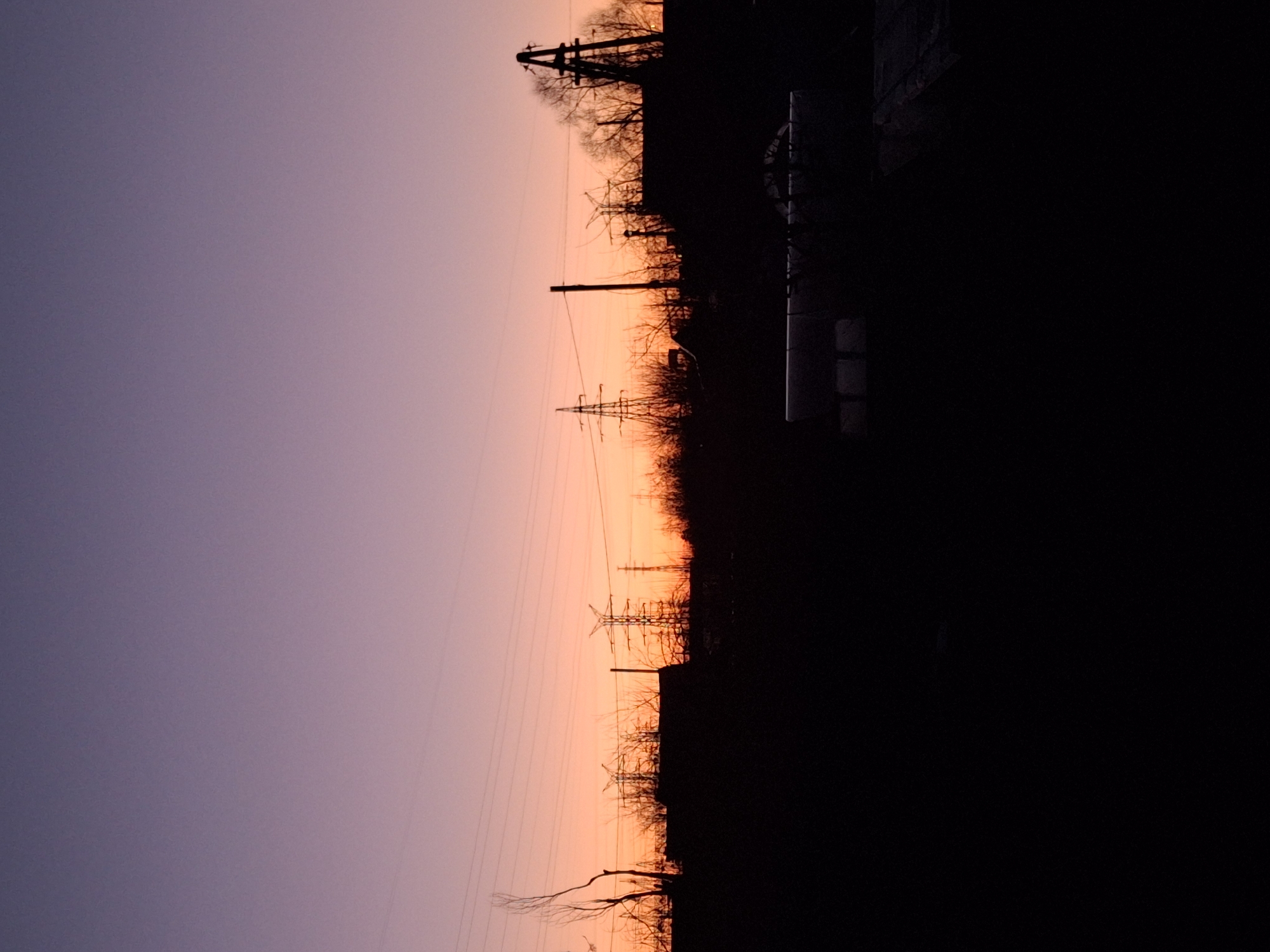 The image captures a sunset in what appears to be the city of Chaikovsky, Russia, as evidenced by the industrial setting and overhead power lines, which are typical of urban and industrial areas. The sky transitions from a purple hue on the left to a warm orange glow on the right, indicating the direction of the setting sun. The silhouette of trees and industrial infrastructure, including power lines and possibly a building, are visible against the sunset backdrop. The lower portion of the image is dominated by dark shadows, obscuring details of the foreground landscape. There are no people visible in the scene. The focus is on the atmospheric effect and the juxtaposition of natural and man-made elements.