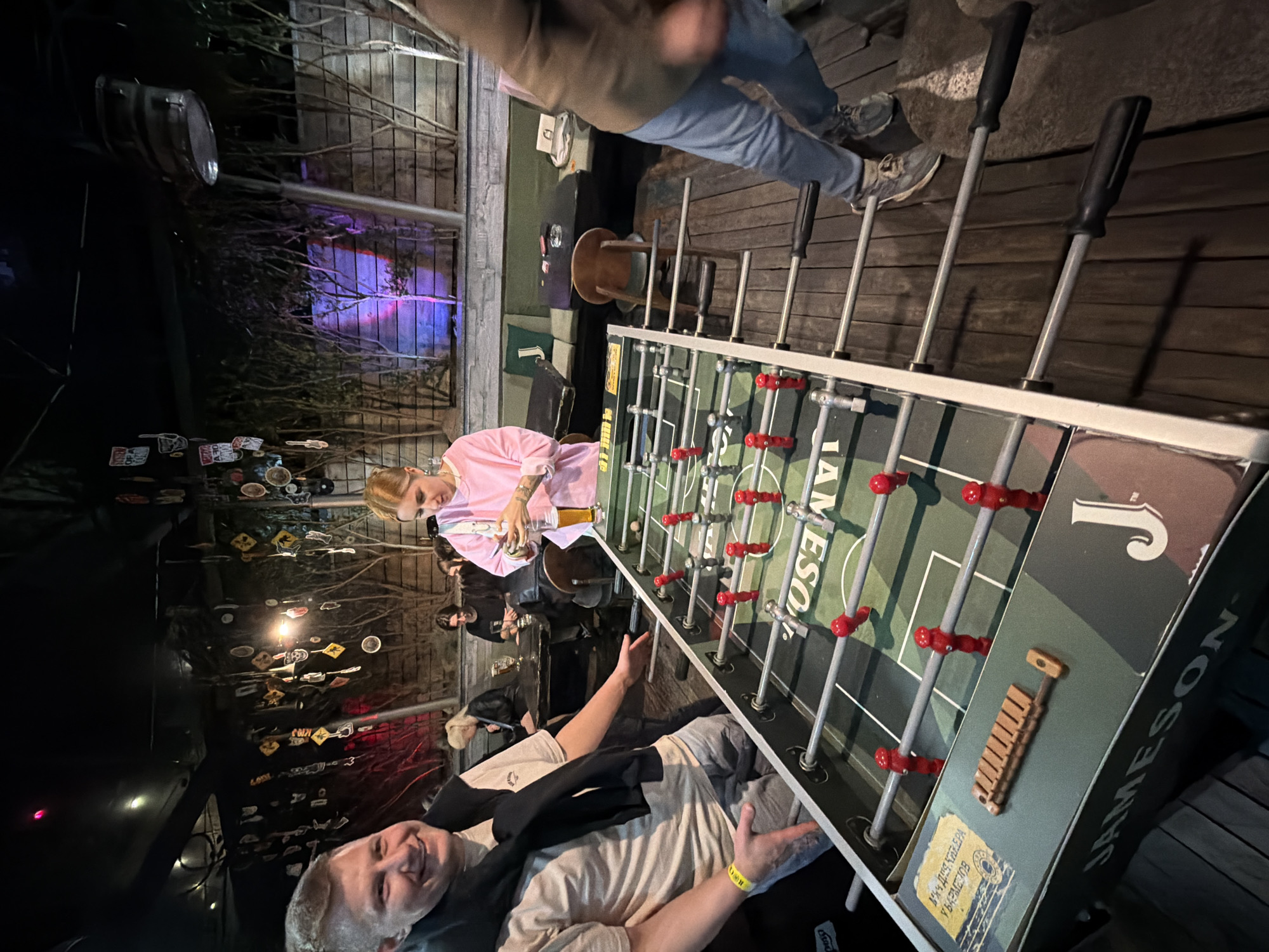 The image depicts a group of people playing foosball at a bar or outdoor venue, possibly in Minsk, Belarus, given the Cyrillic script visible on the foosball table itself. The foosball table is branded "JAMESON." One person is shown actively playing foosball, while another holds a drink and watches. Other individuals are seated nearby. The background includes atmospheric lighting, signage, and various decorations. The scene appears to be a social gathering in a relaxed, outdoor environment.