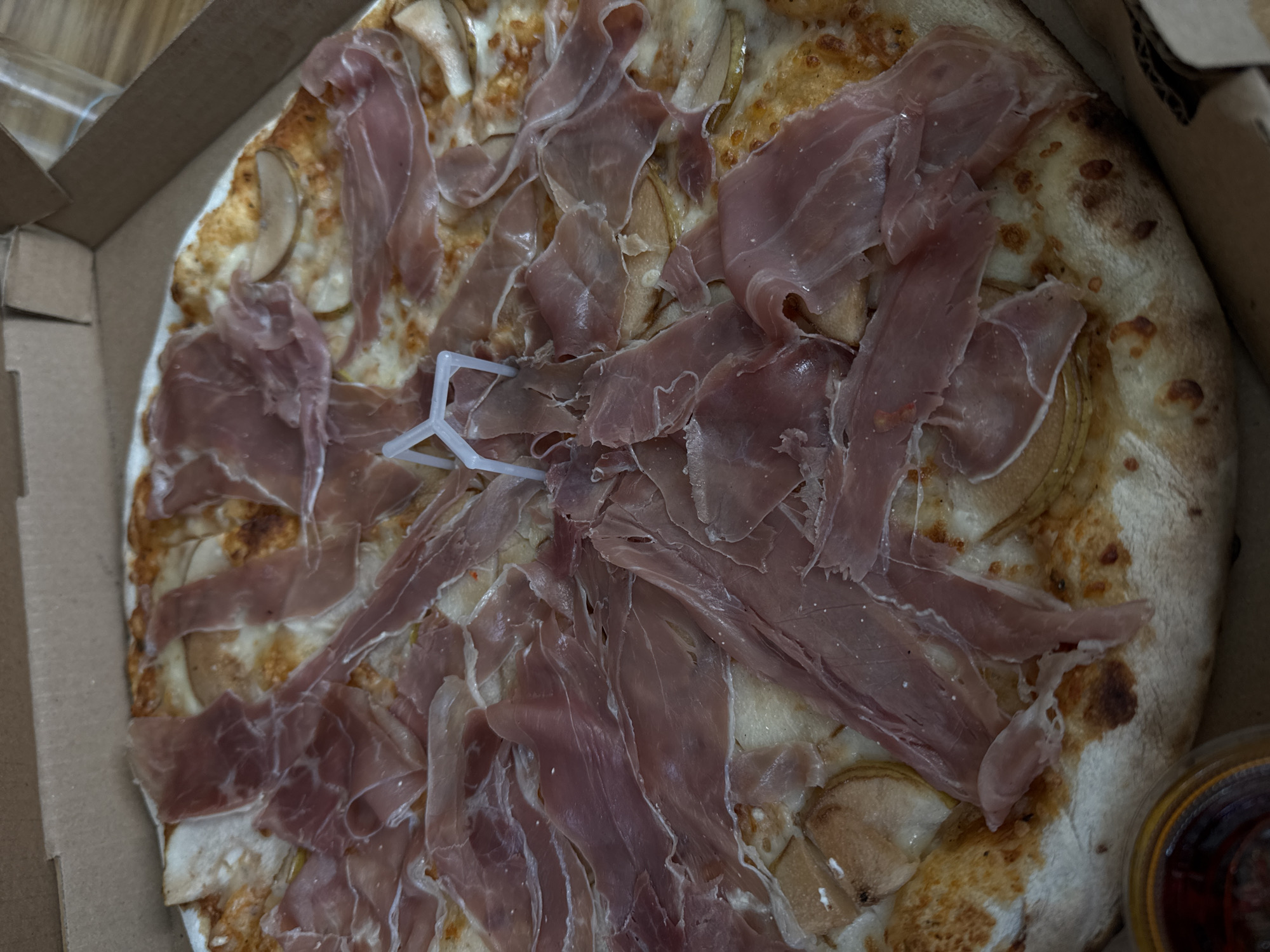 A pizza in a cardboard box is shown from above. The pizza is topped with ham and sliced pears. A white, plastic pizza saver is placed in the center. The scene is likely indoors, perhaps at a restaurant or residence in San Bernardino Tlaxcalancingo, Mexico. A container with a dark fluid is located on the right side, outside of the pizza box. No people are visible.