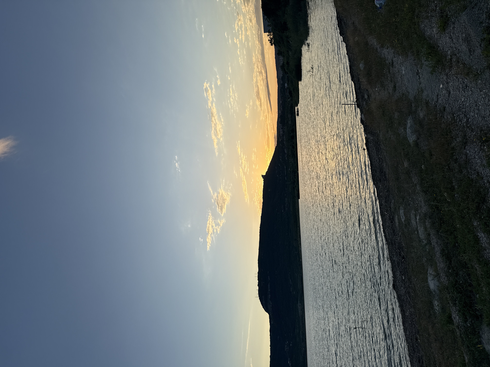 This image depicts a serene lakeside view during sunset, likely taken from the northern bank of Lake Shumen, Bulgaria. The sky transitions from a clear, pale blue at the top to a soft orange and yellow hue near the horizon, indicative of the setting sun. Scattered, wispy clouds catch the fading sunlight, adding texture to the sky.
Dominating the left side of the frame is the silhouette of a prominent hill, topped with several communication towers. This hill is characteristic of the landscape surrounding Lake Shumen, which is part of the Shumen Plateau Nature Park. The dark mass of the hill contrasts sharply with the vibrant sky.
The foreground features the calm, rippling surface of Lake Shumen. The water reflects the warm colors of the sunset, creating a shimmering pathway of light across its expanse. The shoreline in the immediate foreground is composed of rough, dry grass and gravel, with a visible path winding its way along the edge. Sparse vegetation and some scattered debris are also present along the shore. In the distance, on the right side of the image, hints of trees and possibly some buildings can be discerned against the fading light. The overall scene conveys a sense of tranquility and natural beauty at the close of day.