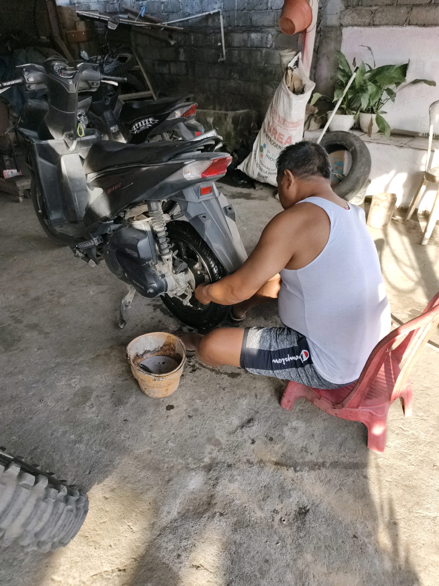 In Tombatu, Indonesia, a man in a white tank top and grey and black Champion shorts is seen cleaning the rear tire of a black scooter. He is kneeling on the concrete floor, with a small, weathered orange bucket containing dirty water beside him. The scooter is parked in what appears to be a covered outdoor space, possibly a garage or workshop, with concrete walls and a corrugated metal roof visible in the background. Various items are scattered around, including a large white bag of what looks like fertilizer, a spare tire, and some potted plants. Sunlight streams in, casting shadows on the floor.